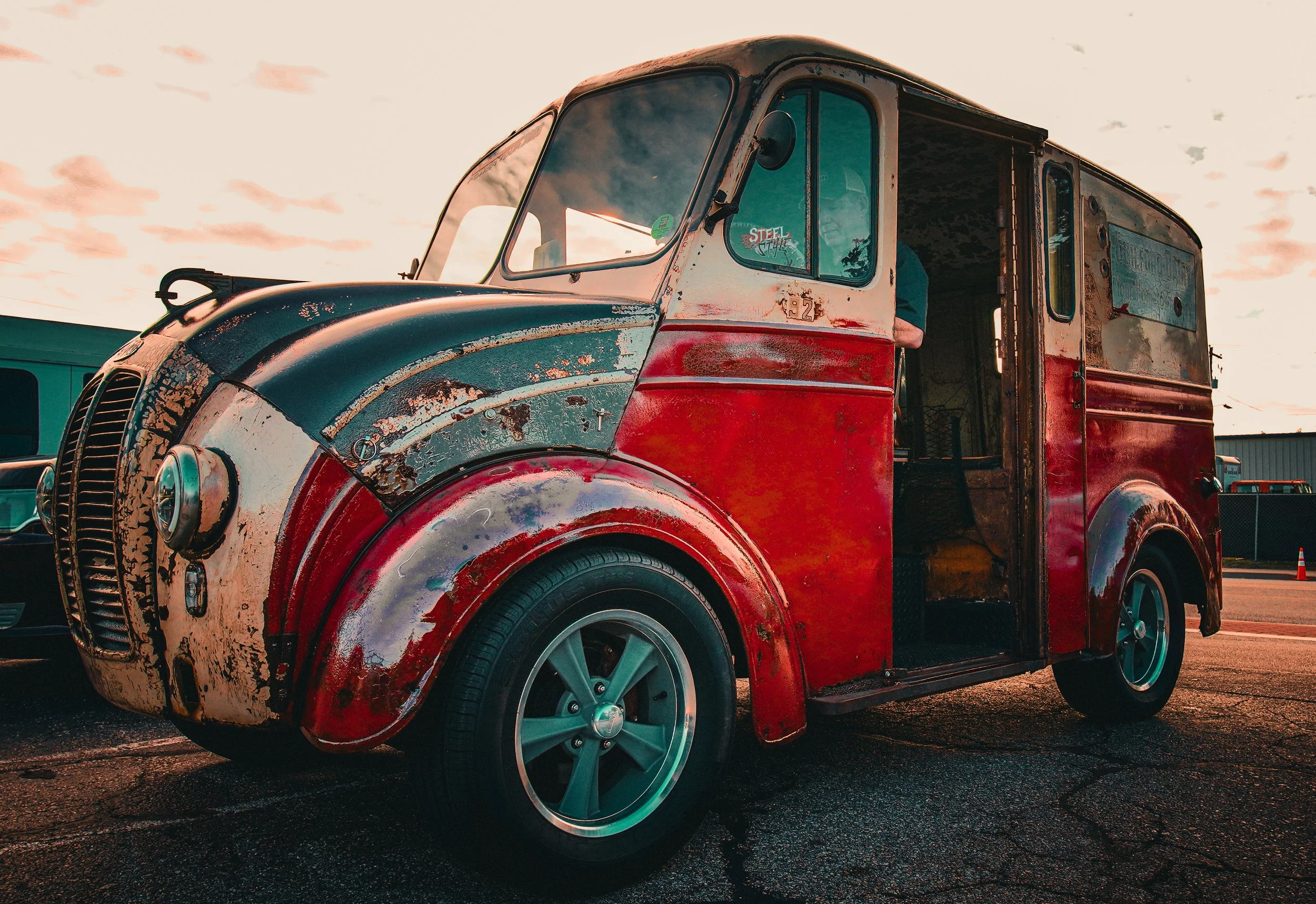 An old, rusted delivery truck with a red and black paint job parked on an asphalt lot at sunset.