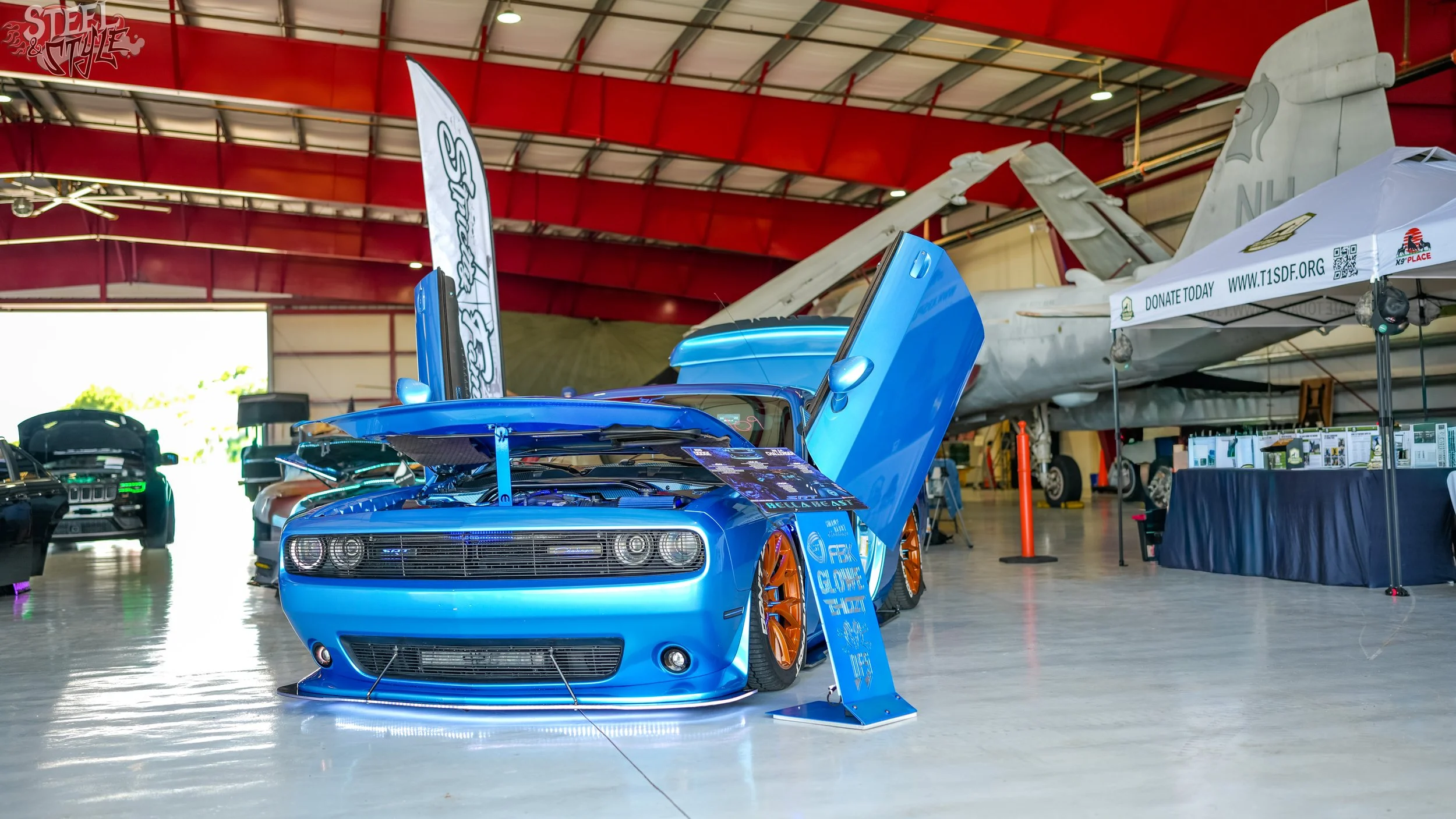 Blue Dodge challenger with car doors open, displayed indoors at an aircraft museum with a large white fighter jet in the background.