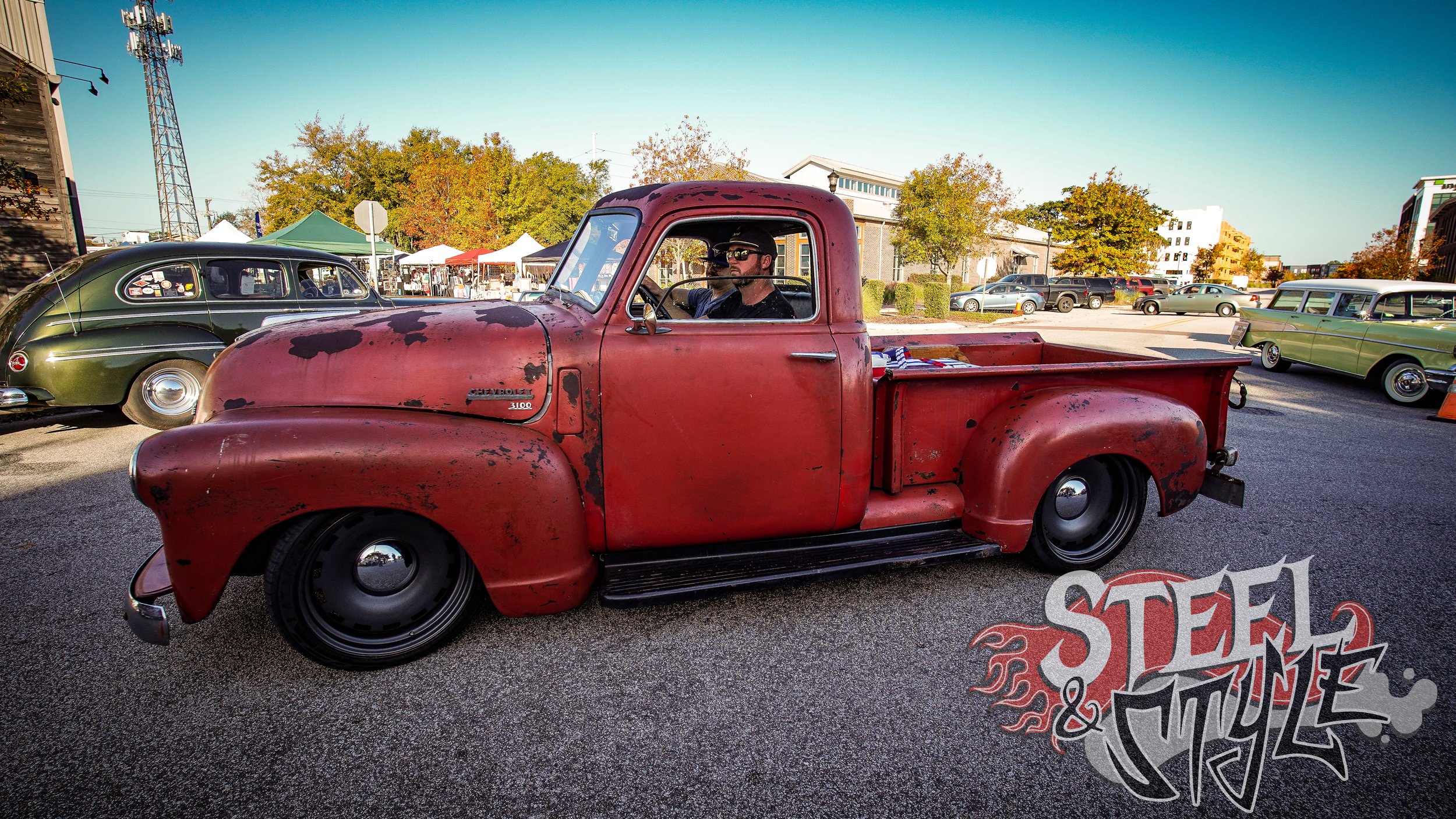 A vintage red Chevrolet pickup truck with a weathered and rusted appearance parked at a car show, surrounded by other classic cars and tents in the background on a sunny day.