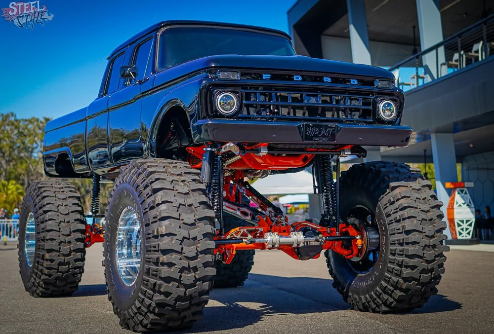 A lifted vintage black Ford truck with oversized off-road tires and red suspension components at an outdoor event.