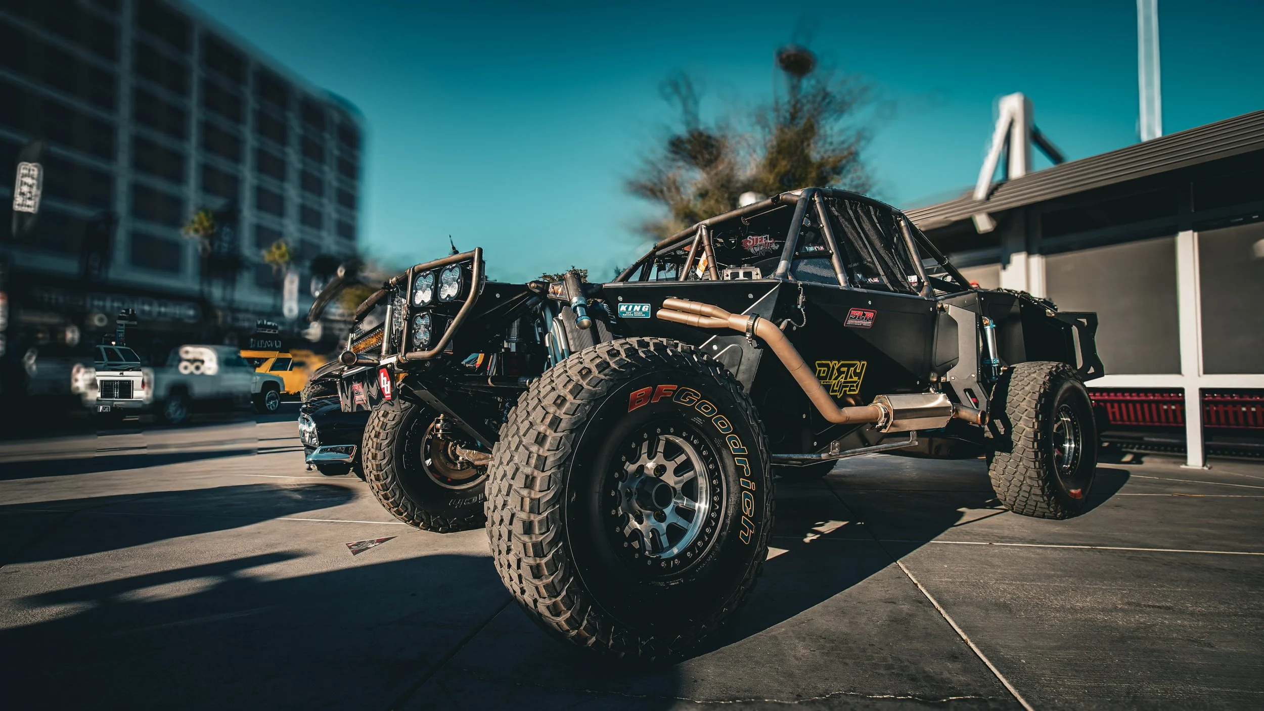 Off-road race car parked outdoors with a cityscape background and clear blue sky.