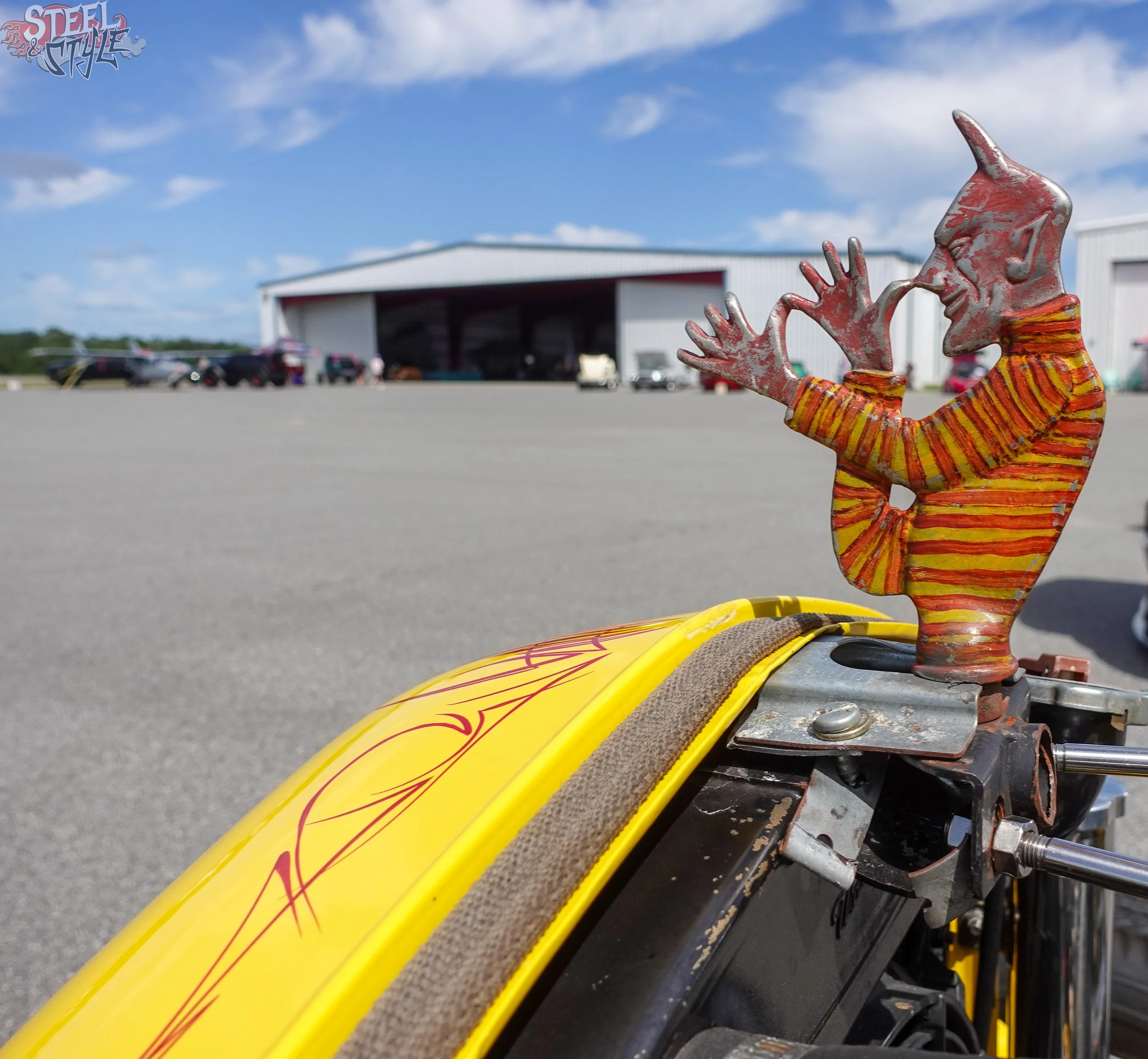 This is a close-up view of a drag racing car's front nose, which is bright yellow with red flame graphics. The image shows the car's frame, and there's a small decorative metal sculpture of a person wearing a hat with a musical note, painted in weath
