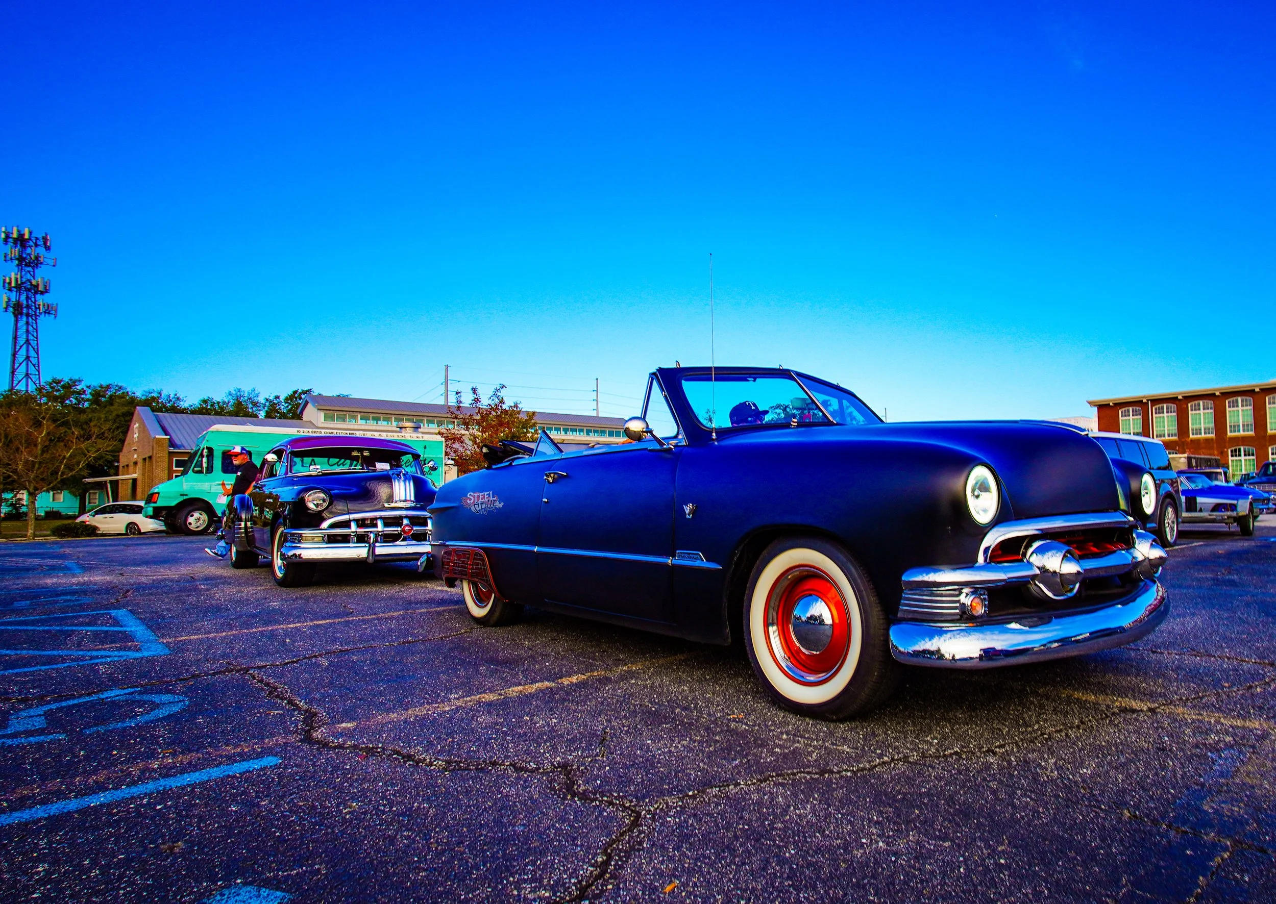 Vintage cars parked in a lot during daytime with clear blue sky, people walking around, and buildings in the background.