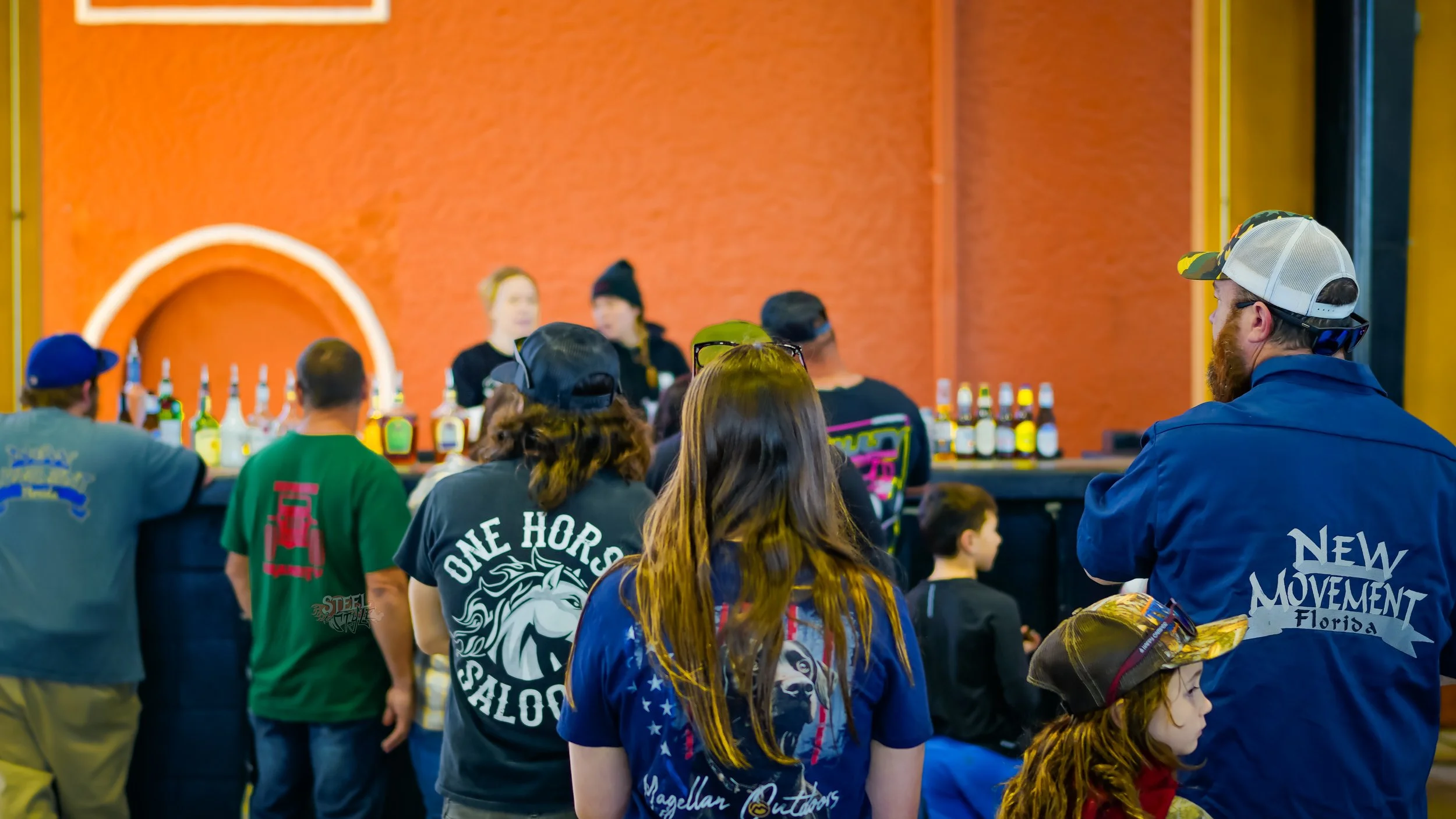 Group of people gathering at a bar with a row of bottled drinks, two women behind the bar, and a large orange wall in the background.