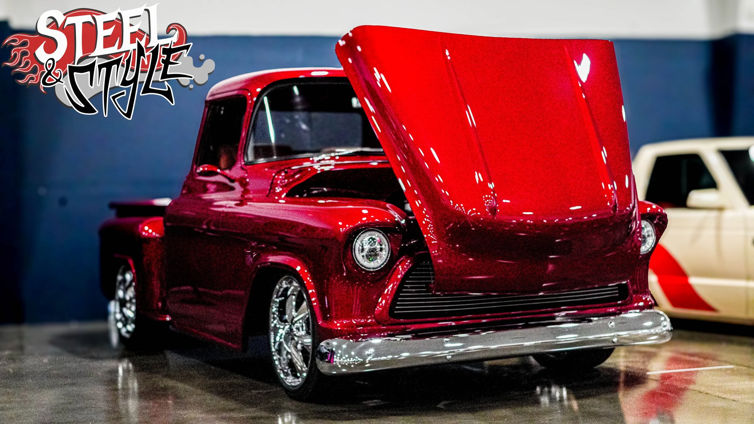 A red vintage pickup truck with a raised hood at an auto show, with a logo reading 'Steel & Style' in the top left corner.