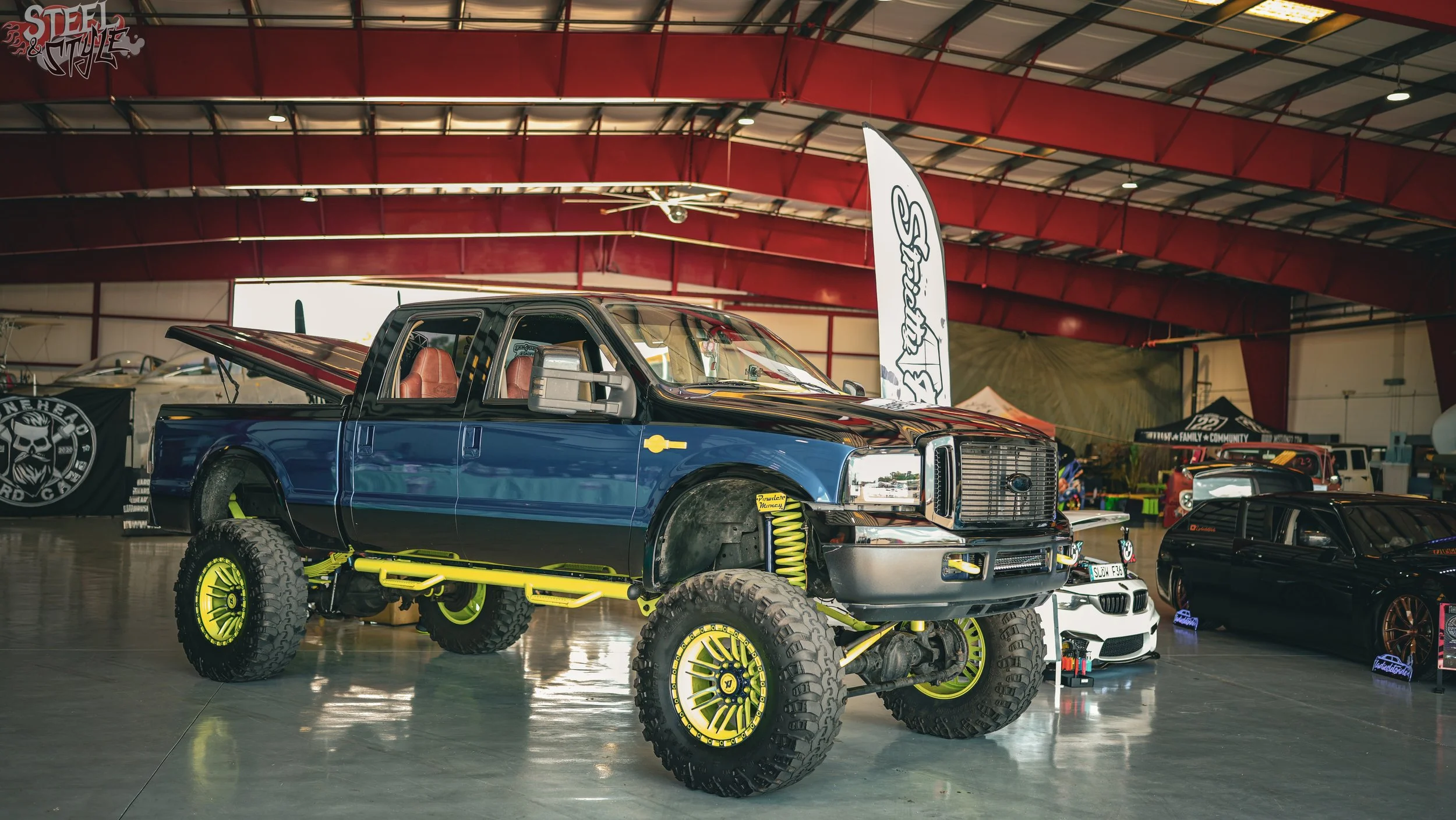An indoor car show featuring a modified off-road truck with large tires and bright yellow suspension components, surrounded by other customized vehicles in a spacious exhibition hall.