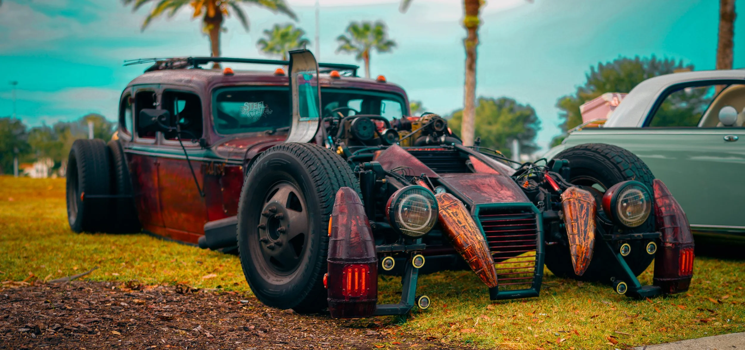 A vintage car with its body removed, exposing the engine and internal components, parked on a grassy area with palm trees in the background.