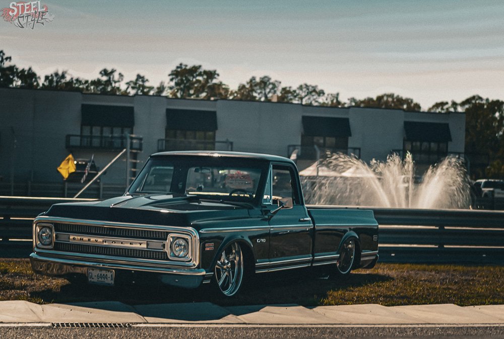 A vintage black and white Chevrolet C10 pickup truck parked on a grassy area next to a racing track with a fountain in the background.