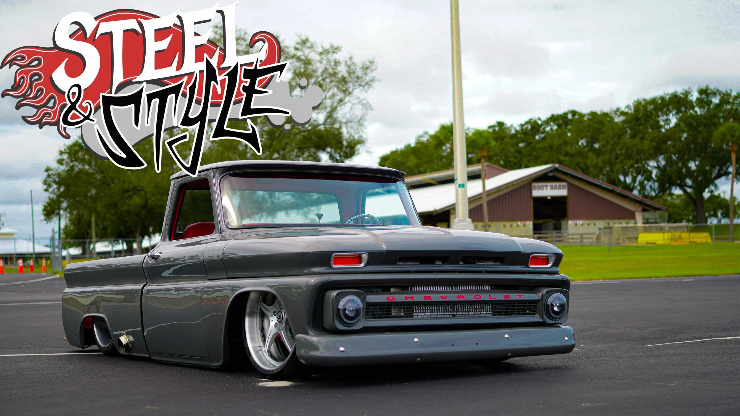 A customized gray vintage Chevrolet pickup truck parked on a paved surface with lowered suspension and large wheels. There is a stylized graffiti text overlay reading 'Steel & Style' in the top left corner, and buildings and trees in the background.