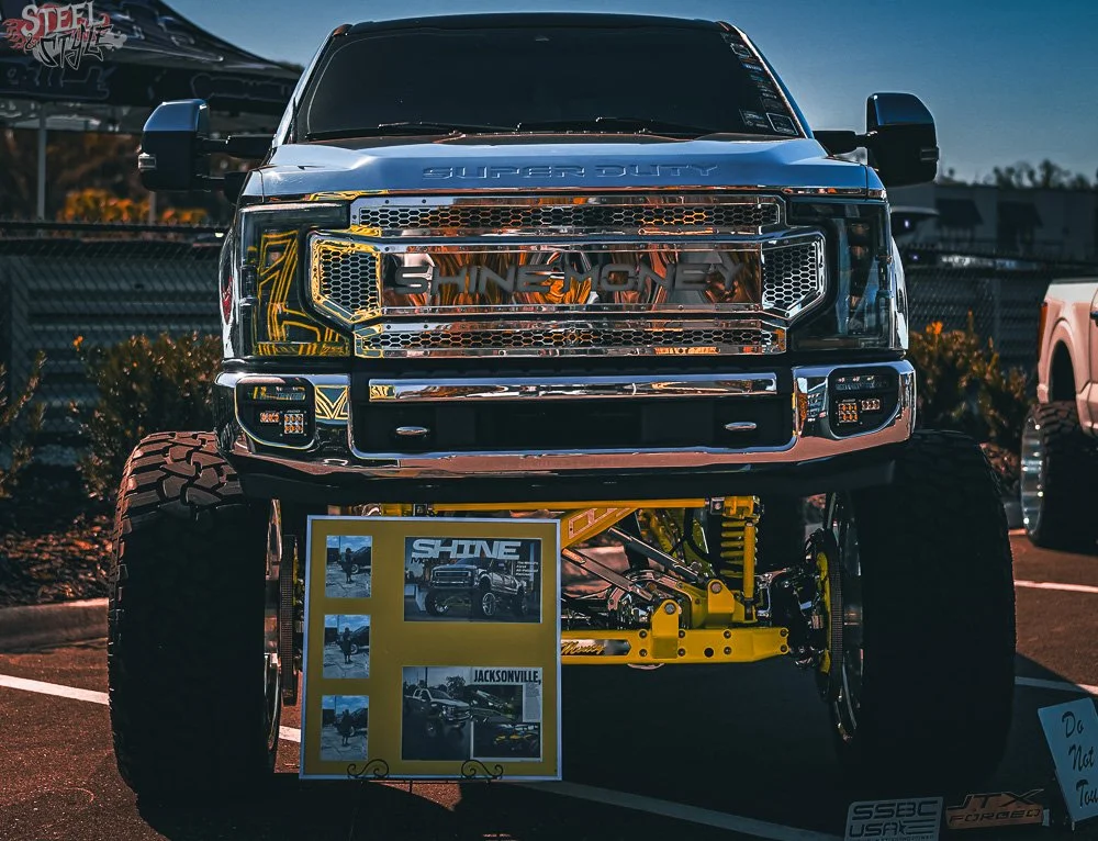 Front view of a highly modified, large yellow monster truck with oversized tires, chrome grill, and reflective details, on display at an outdoor event during sunset.