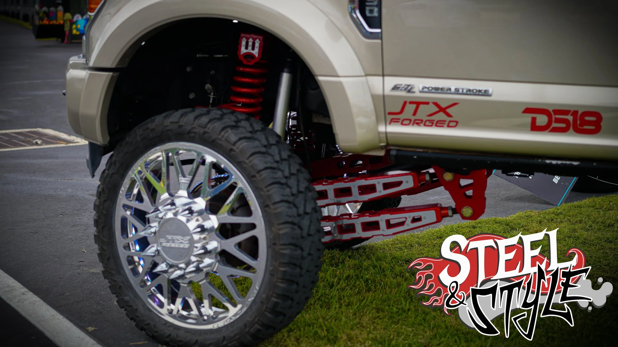 Close-up of a modified off-road vehicle showing its lifted suspension, large tire, and chrome wheel. The vehicle has stickers reading 'JTX Forged,' 'D518,' and 'STEEL & CYCLE' in graffiti-style lettering.