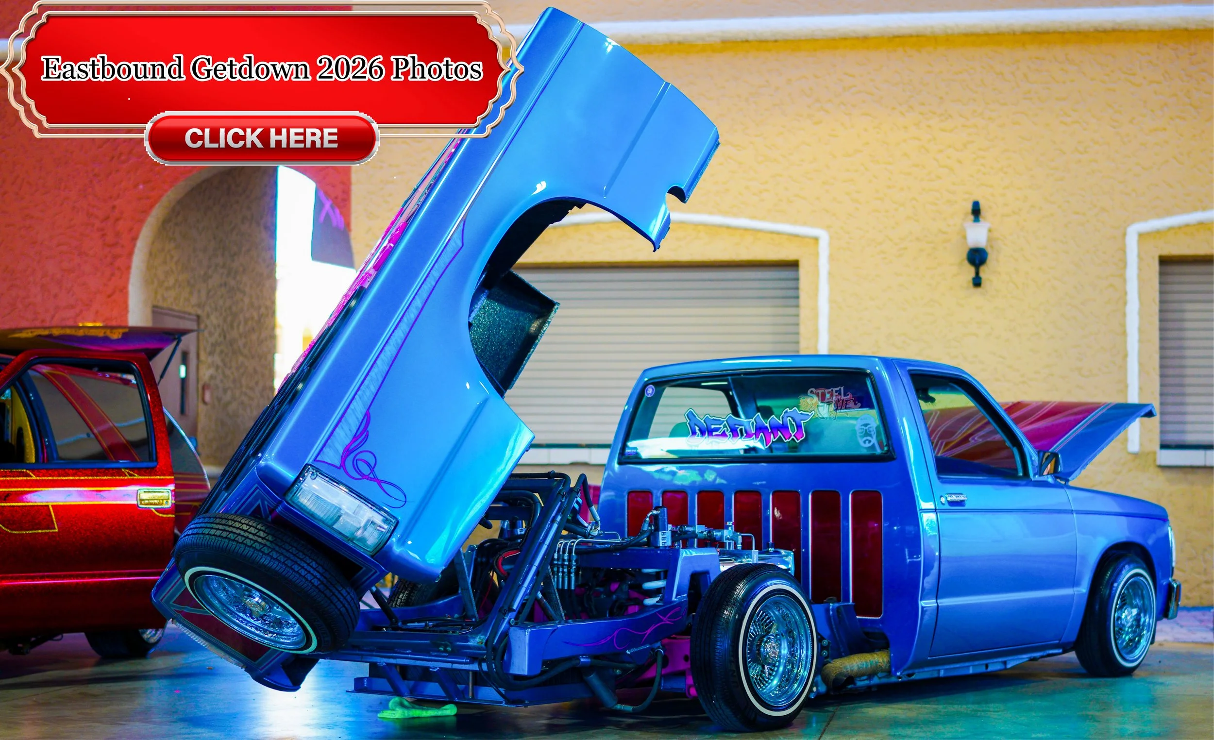 A customized vintage pickup truck with an elevated hood exposing its engine, painted in shiny blue with purple flame decals, parked indoors near a yellow wall with a window and door, alongside a red vintage car.