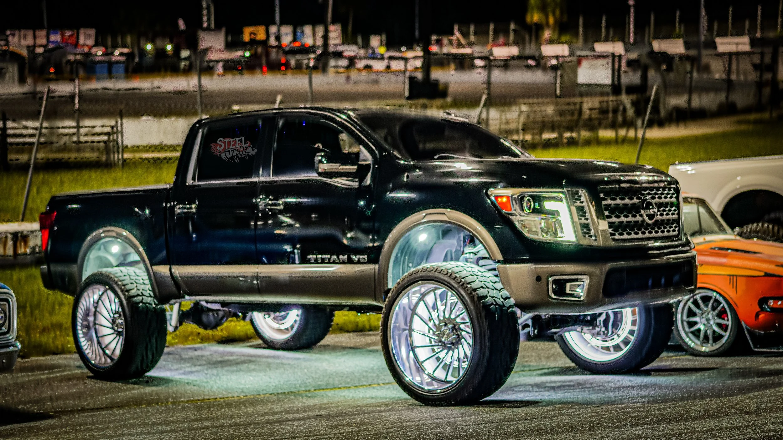 A black Nissan Titan pickup truck with illuminated, oversized wheels and tires parked at night in a lot with other vehicles, including an orange sports car nearby.