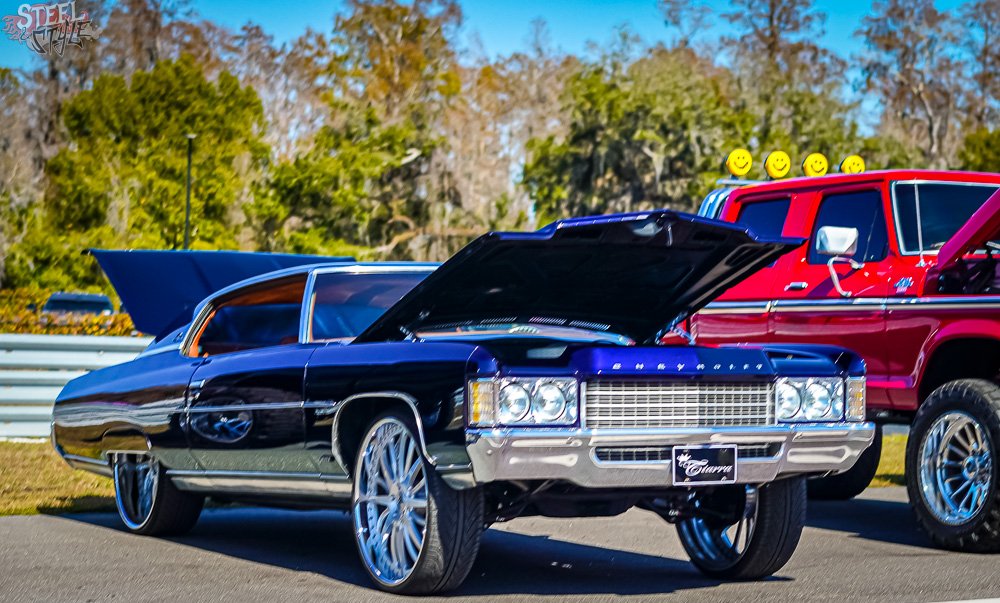 A vintage black Cadillac with chrome accents and large aftermarket wheels, parked outdoors with its hood open. In the background, a red pickup truck with smiley face lights on top of the cab. Some trees and blue sky are visible.