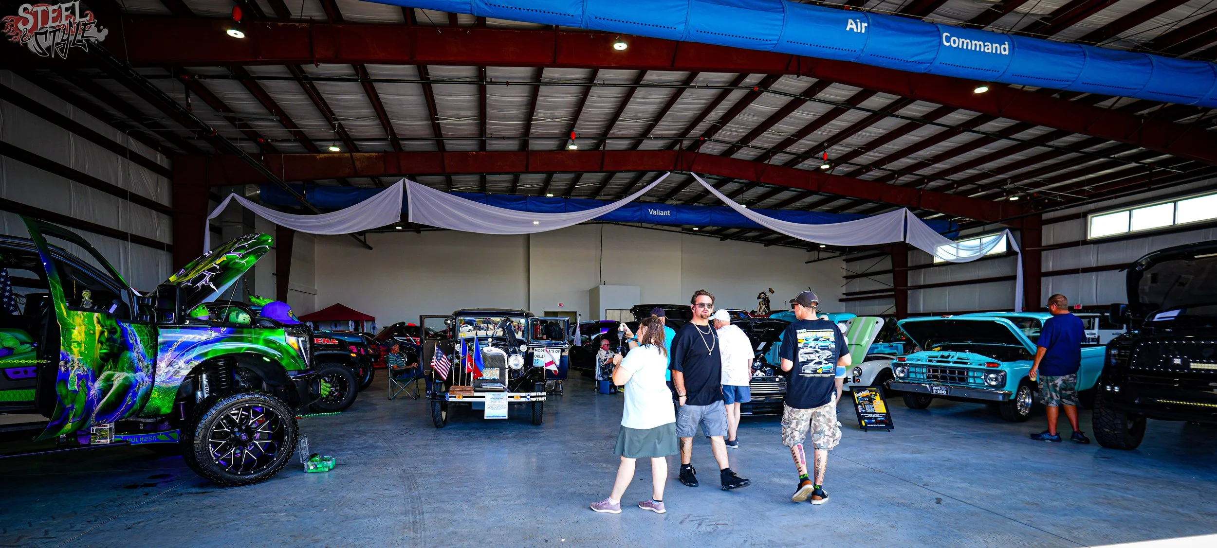 Inside a large exhibition hall with various vintage and custom cars on display, including a colorful truck and a classic black car. Several people are walking around and observing the cars. The hall has a high ceiling with hanging banners and drapery