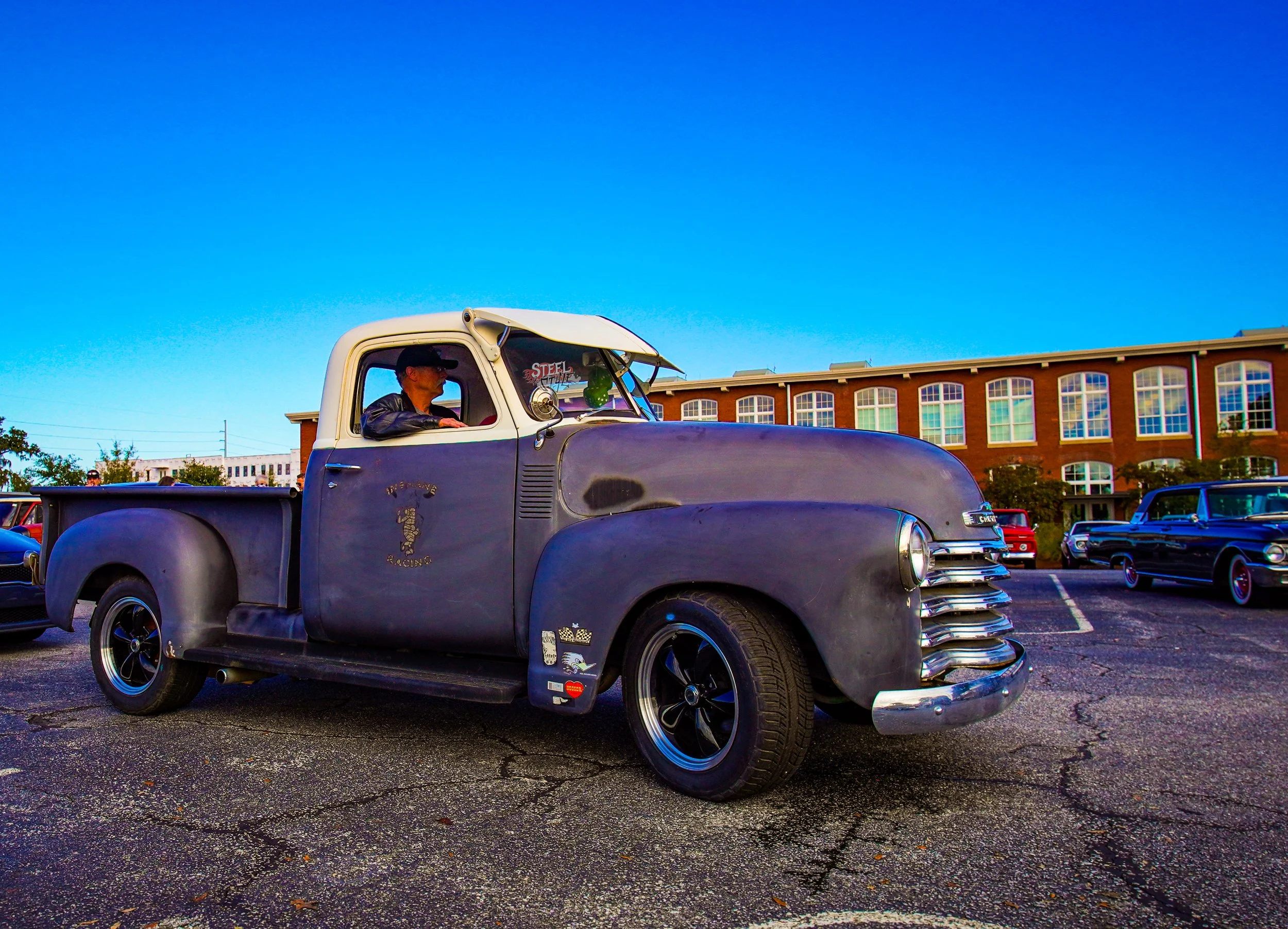Vintage pickup truck parked in an outdoor lot with other classic cars, a modern building in the background, and a bright blue sky.
