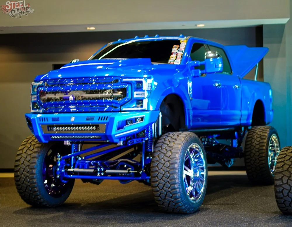 A blue monster truck with oversized tires and a lifted suspension on display at an auto show.