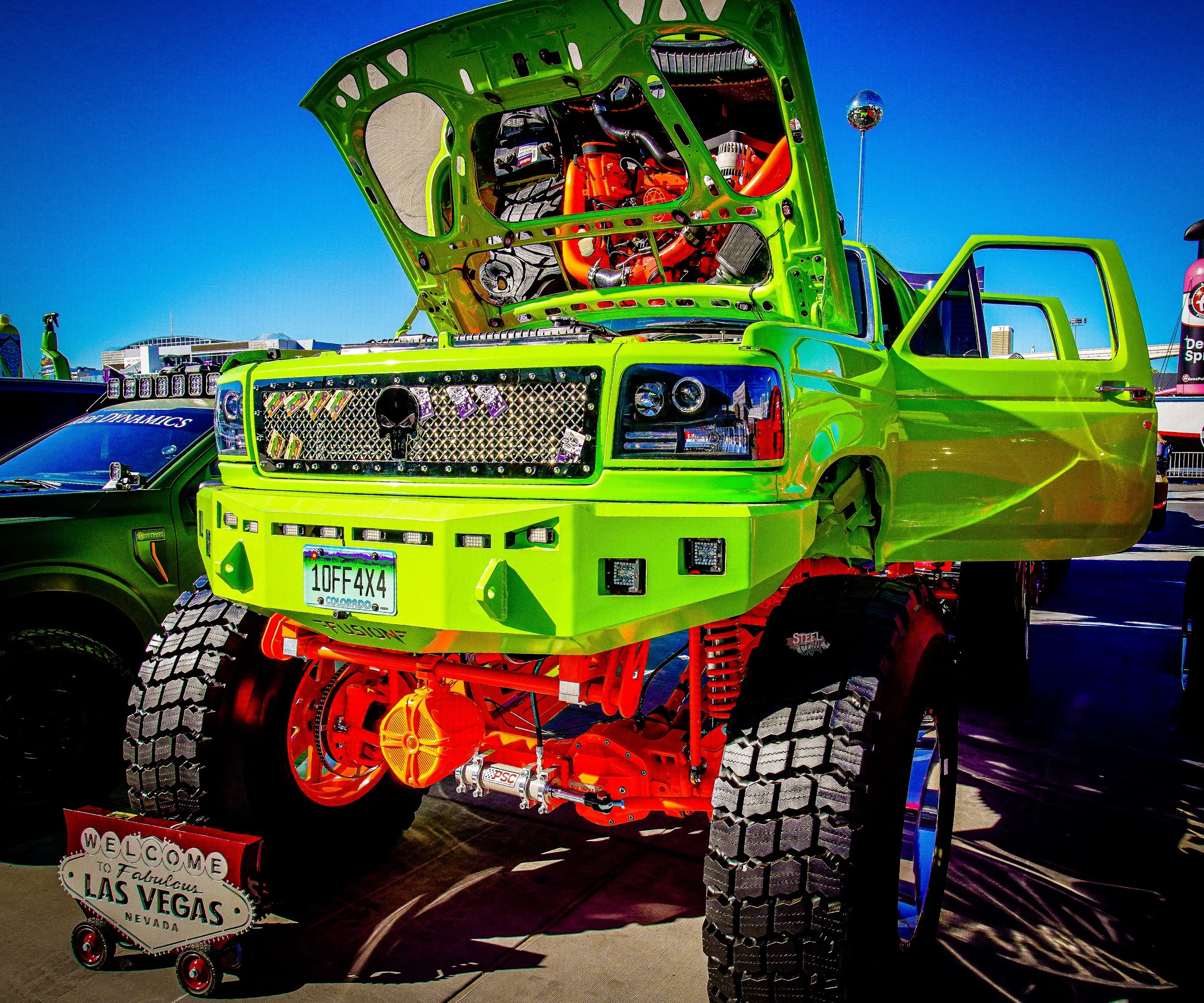 Bright green custom-built off-road vehicle with exposed engine, large tires, and a Nevada license plate, parked outdoors at a car show with a blue sky and other vehicles around.