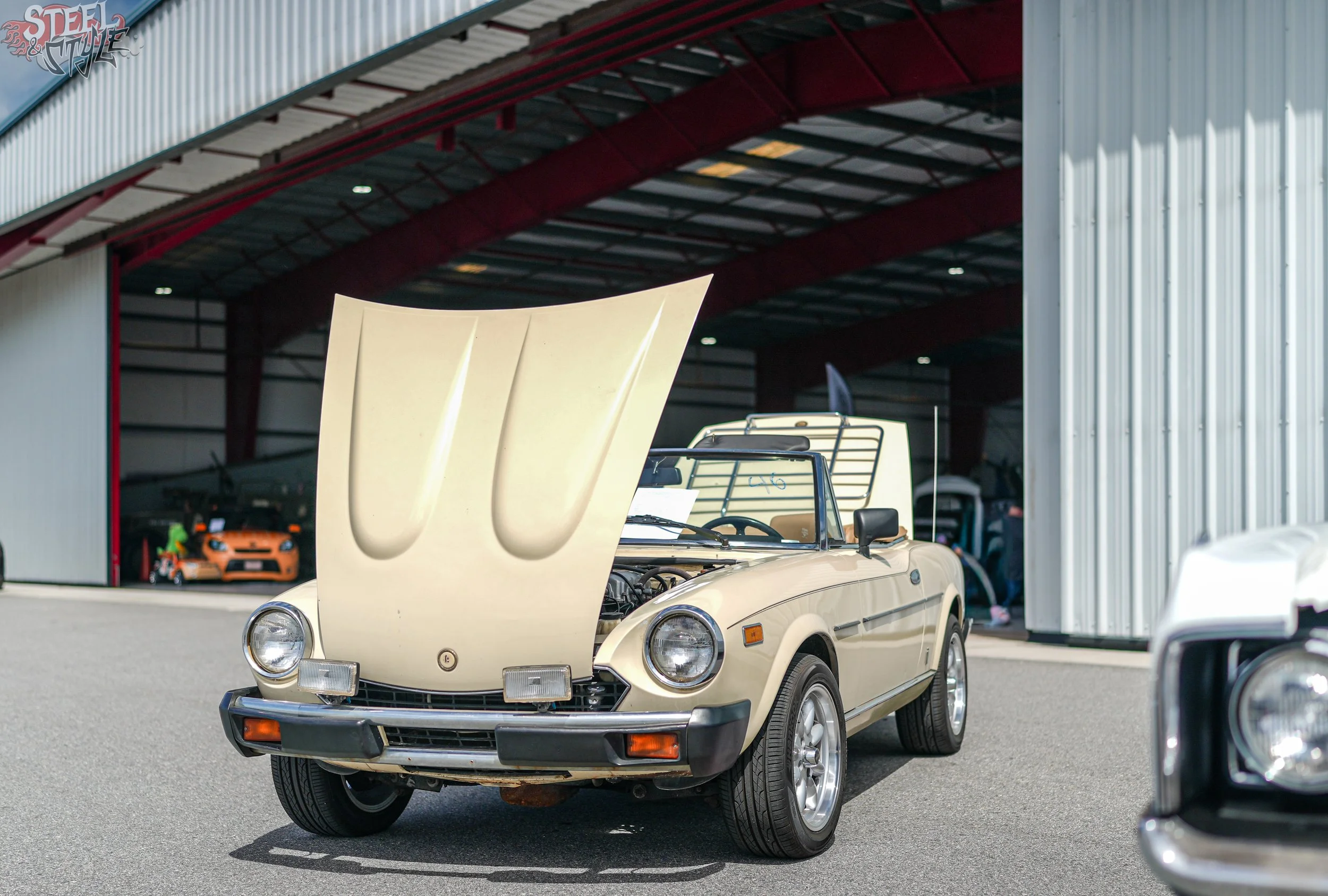A vintage beige convertible car with its hood open, parked outside a building with a red and gray exterior, and other cars visible in the background.