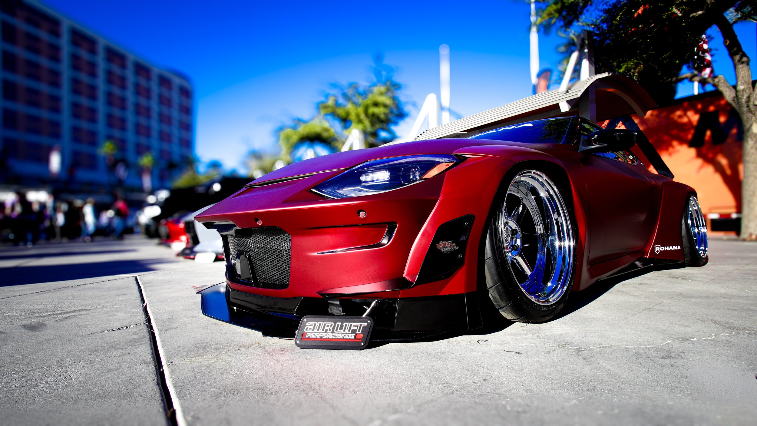 Red modified sports car with large wheels parked on a city street during a car show.