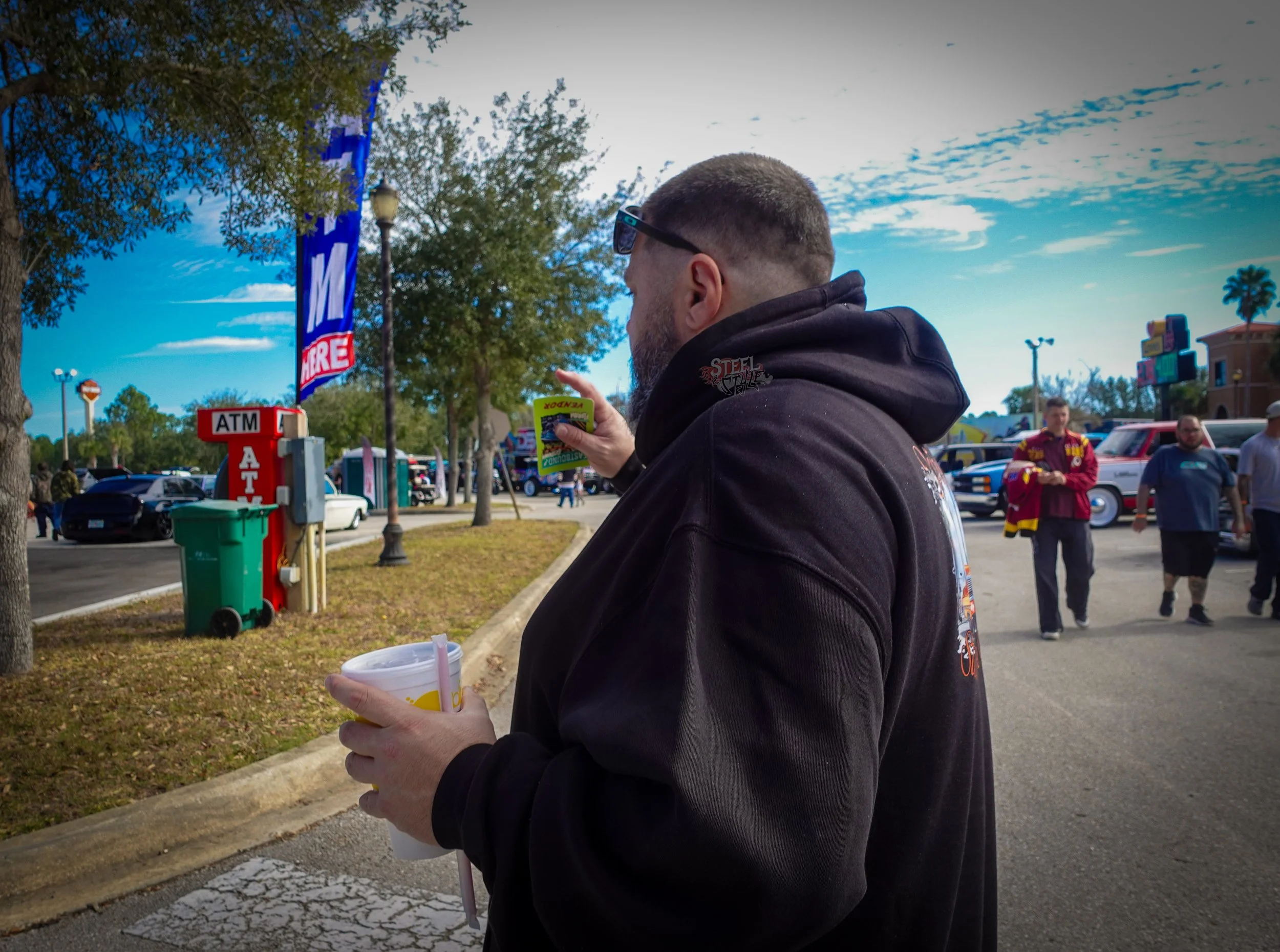 A man wearing sunglasses and a black hoodie, holding a drink in his left hand and reading something on a yellow card with his right hand, standing in a parking lot with trees and a blue sky in the background.