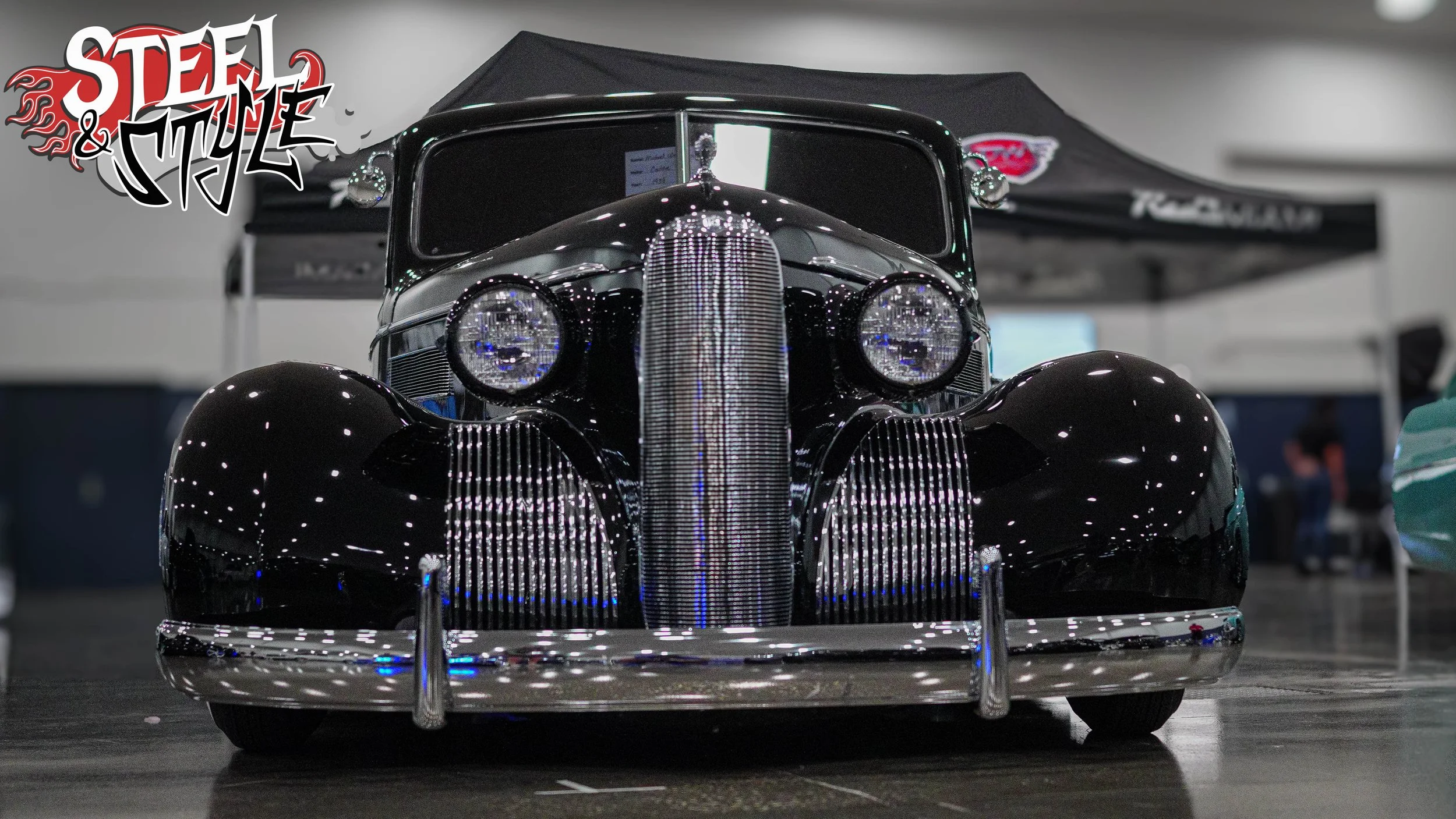 A vintage black automobile with a shiny exterior and prominent chrome details, displayed at an indoor car show. The car has round headlights, a curved grille, and a sleek, rounded body style.