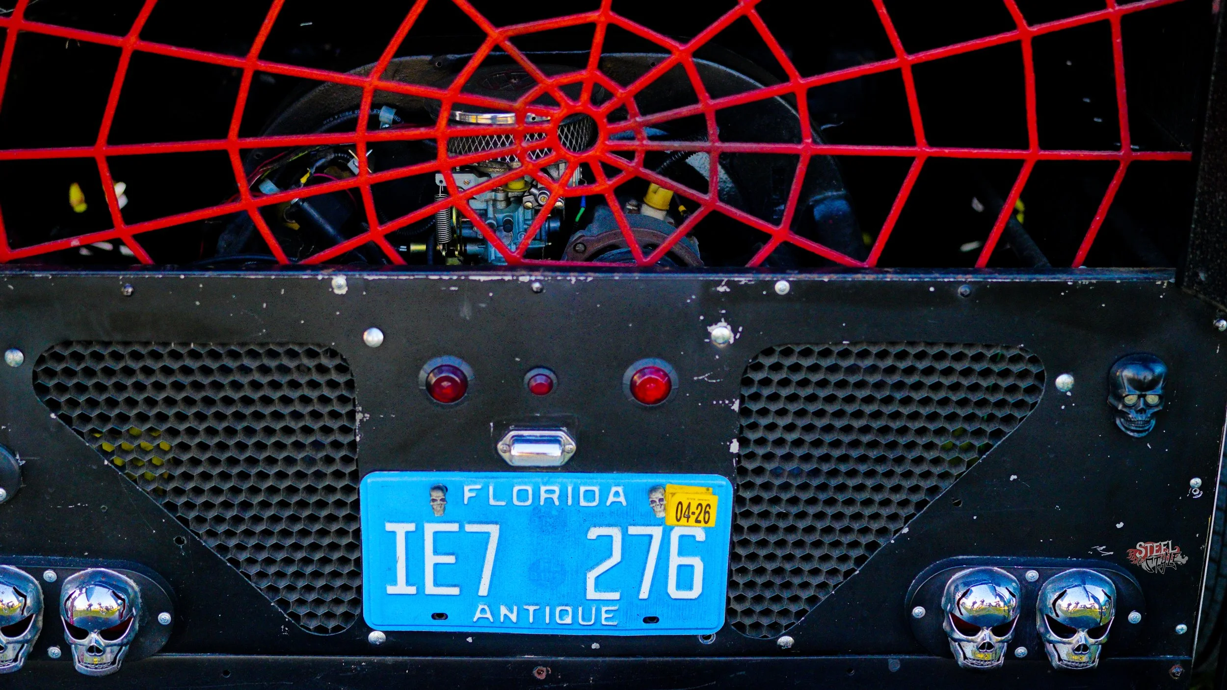 The back of a custom vehicle with a Florida license plate reading 'IE7 276' labeled as 'ANTIQUE', decorated with skull ornaments, featuring a red protective grille and visible mechanical parts behind it.