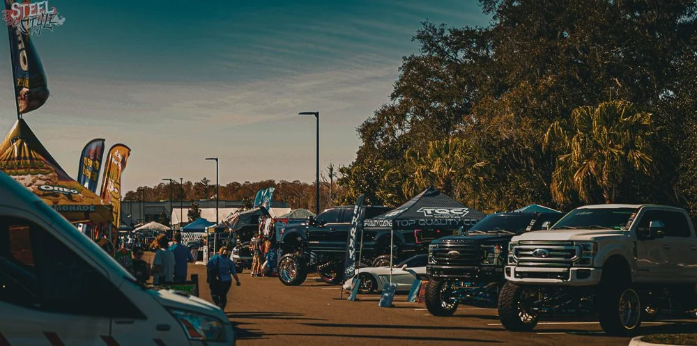 Outdoor car show featuring trucks, tents, and flags on a sunny day with trees in the background.