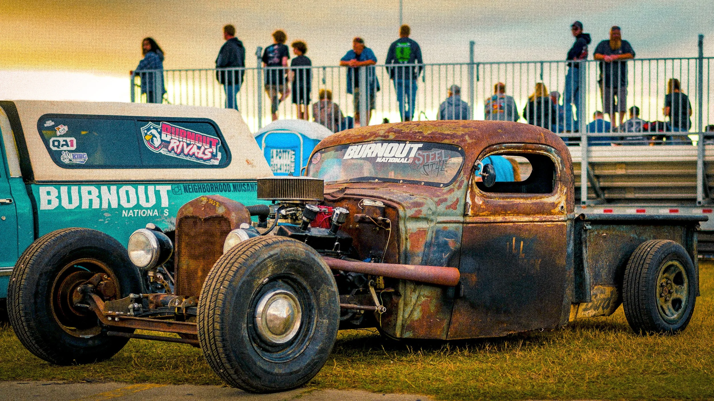 Old, rusty pickup truck parked on grass at a car show, with spectators standing on a second-story viewing platform in the background.