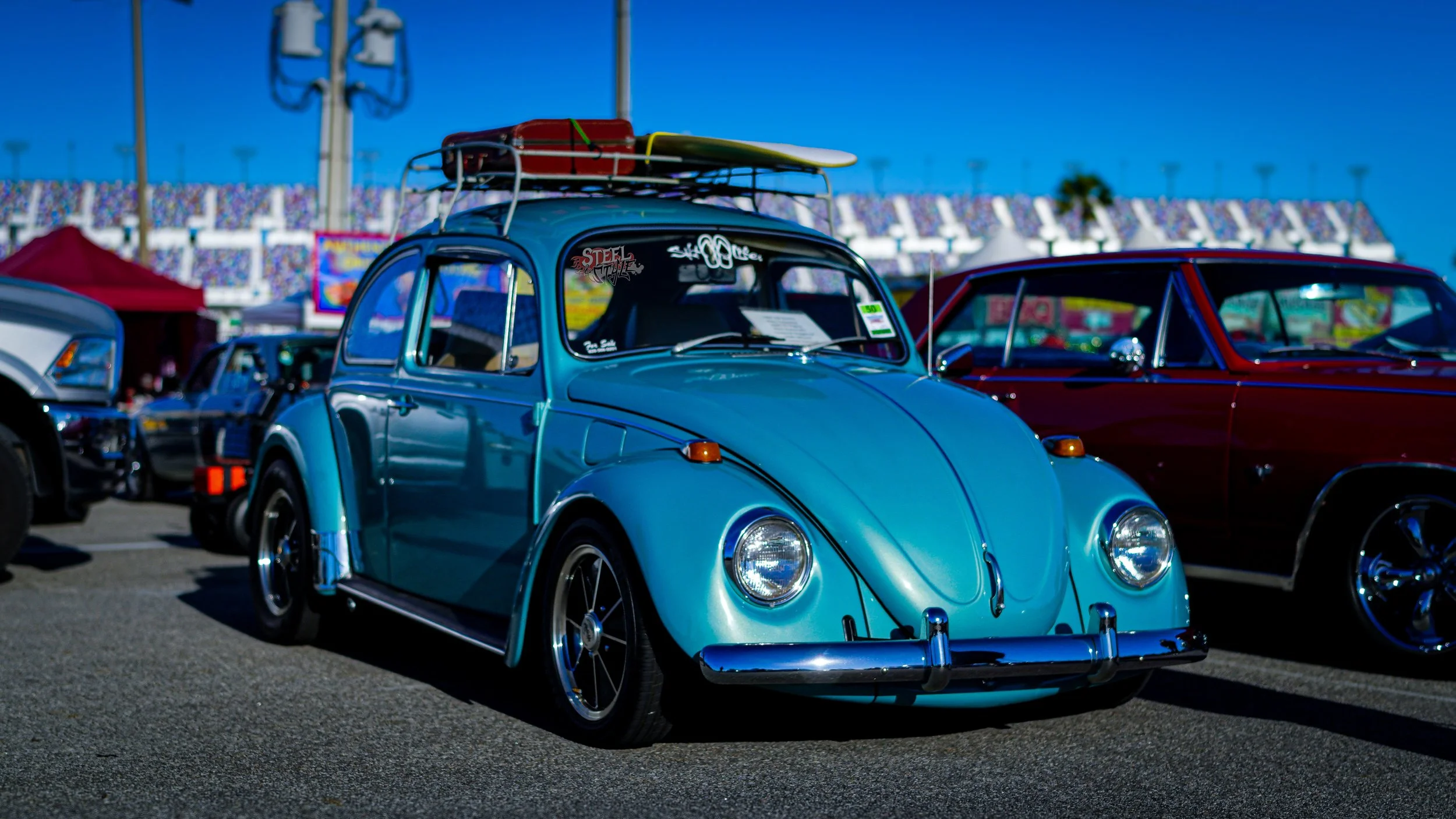 A vintage blue Volkswagen Beetle with a surfboard on the roof, parked at a car show with other classic cars in the background.