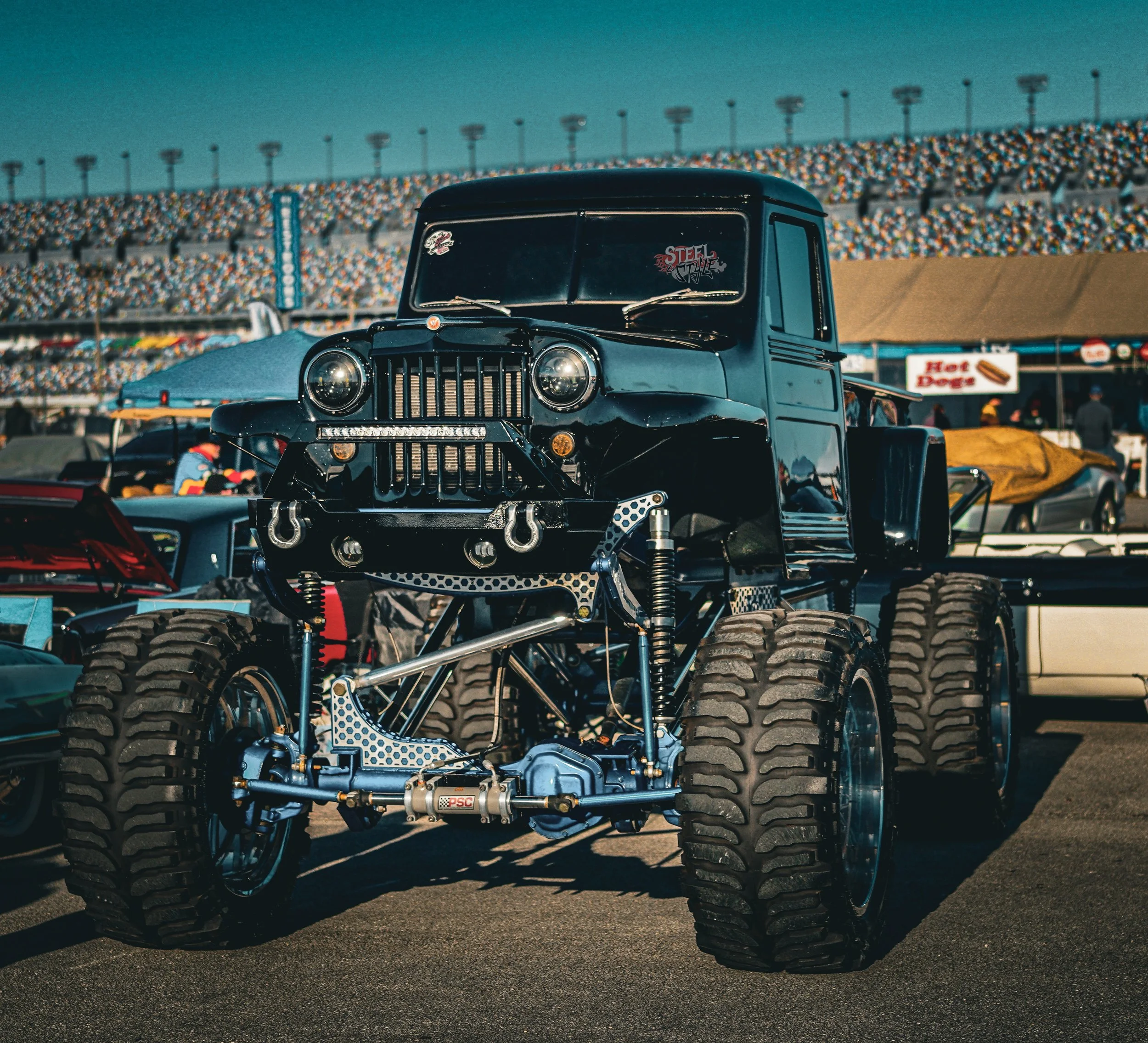 A custom-built off-road truck with oversized tires displayed at a car show, with a racetrack and audience in the background.