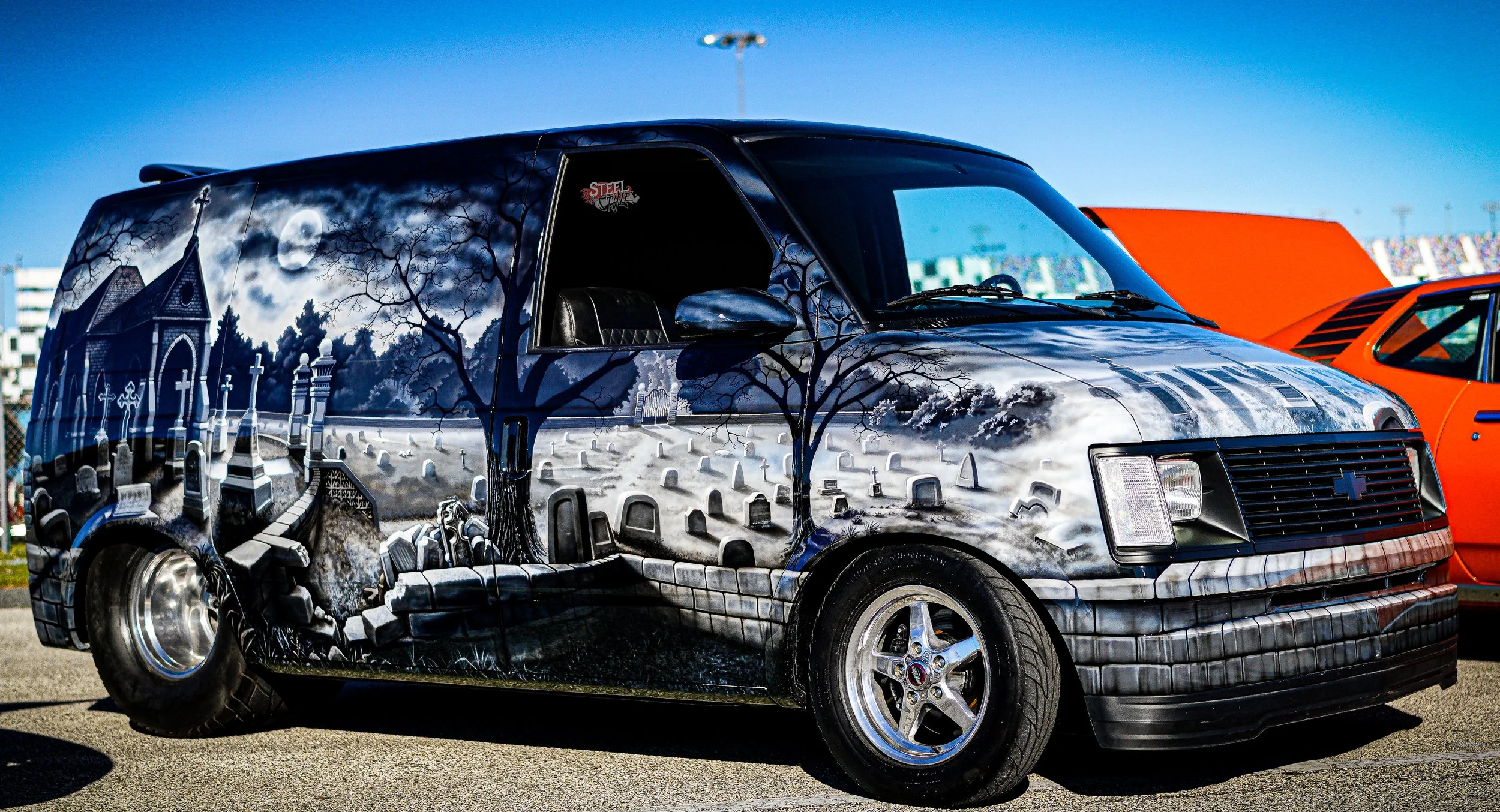 A van with cemetery and spooky night scene painted on its side, featuring a full moon, tombstones, a church, trees, and a stone path.