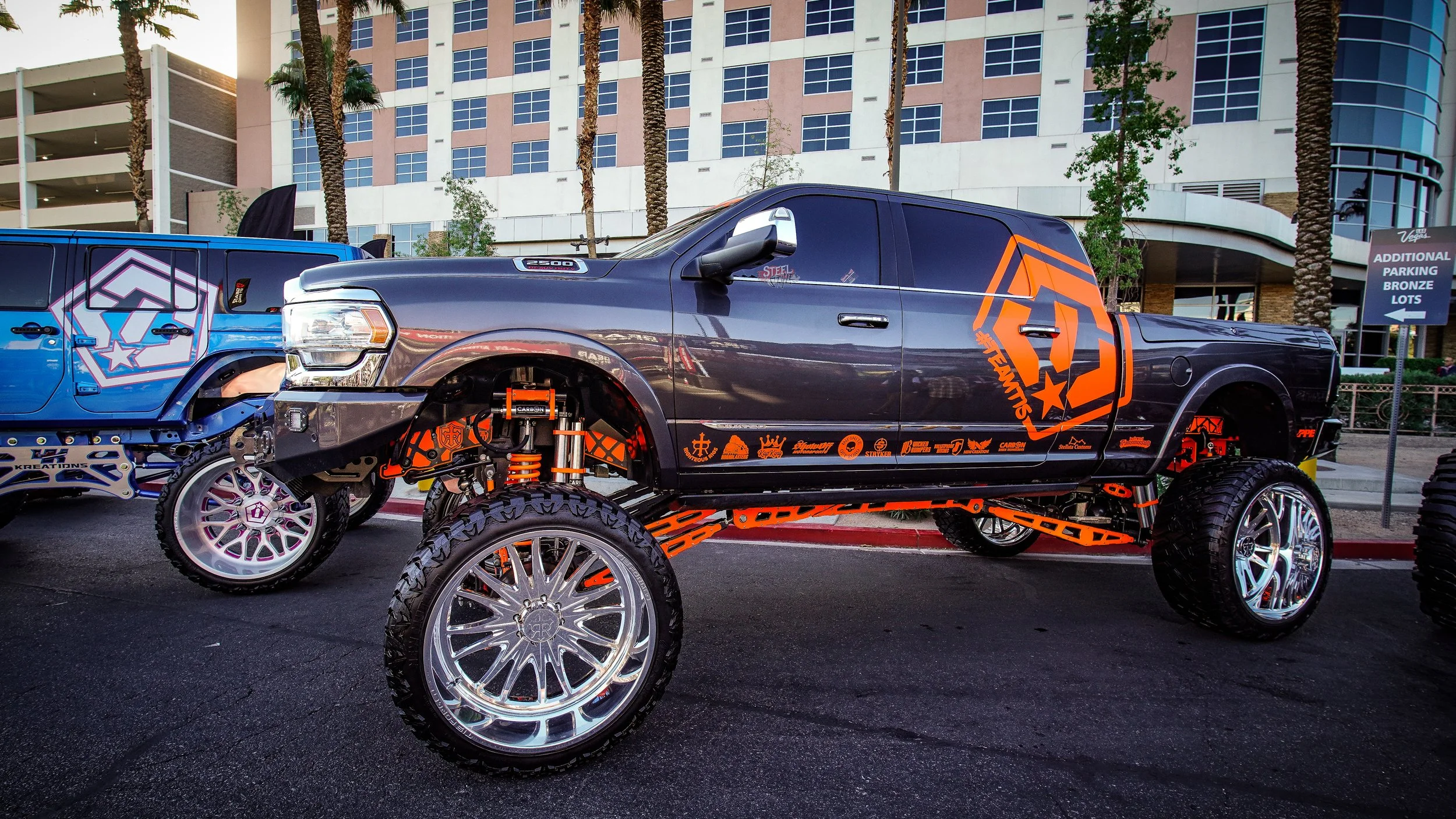 A heavily modified pickup truck with large chrome wheels and orange suspension components, parked outdoors with a modern building and palm trees in the background.
