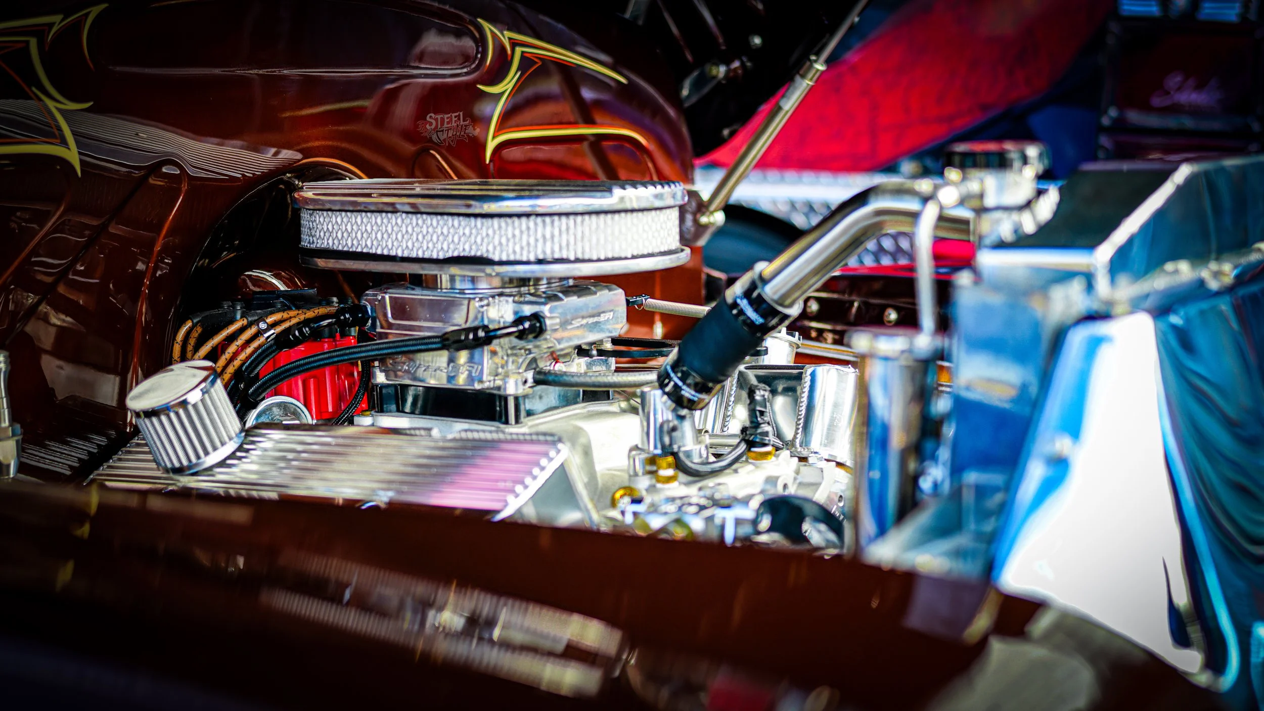 Close-up of a vintage race car engine with shiny metallic components, red distributor cap, and black hoses, with a brown hood and colorful interior in the background.
