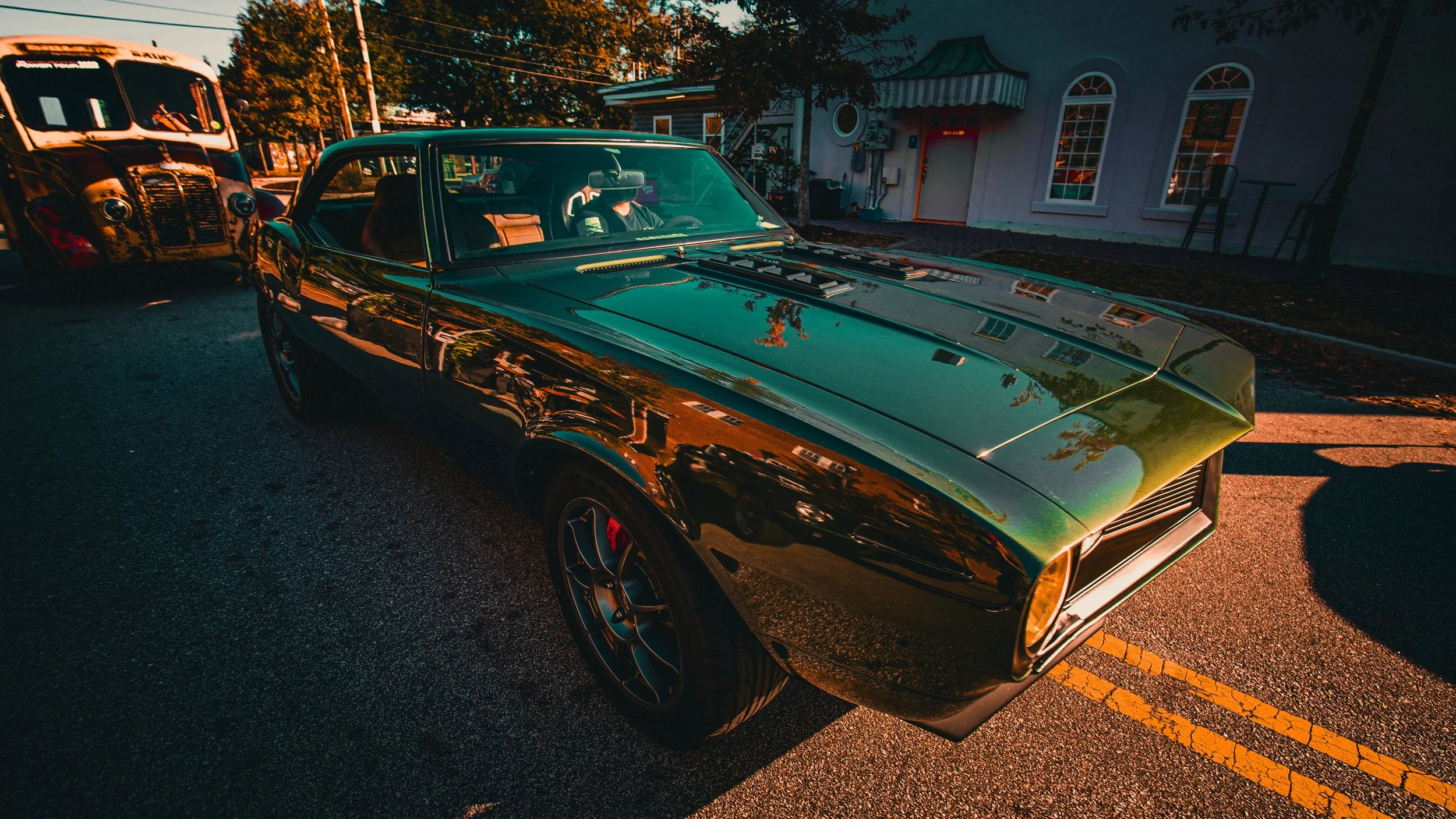 A shiny black vintage muscle car parked on the street with a classic bus in the background. The car's surface reflects the surrounding trees and buildings, and a person is visible inside the vehicle.