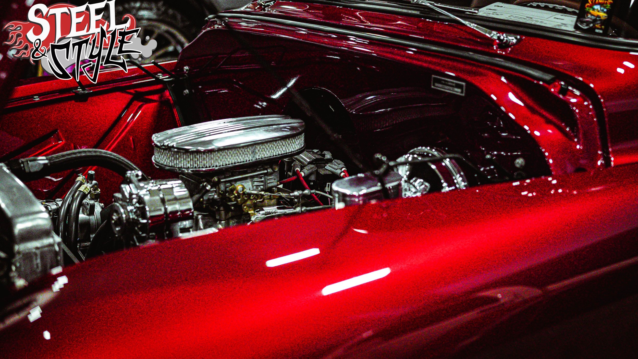 Close-up view of a vintage car engine inside a shiny red car with a 'Steel & Style' logo in the top left corner.