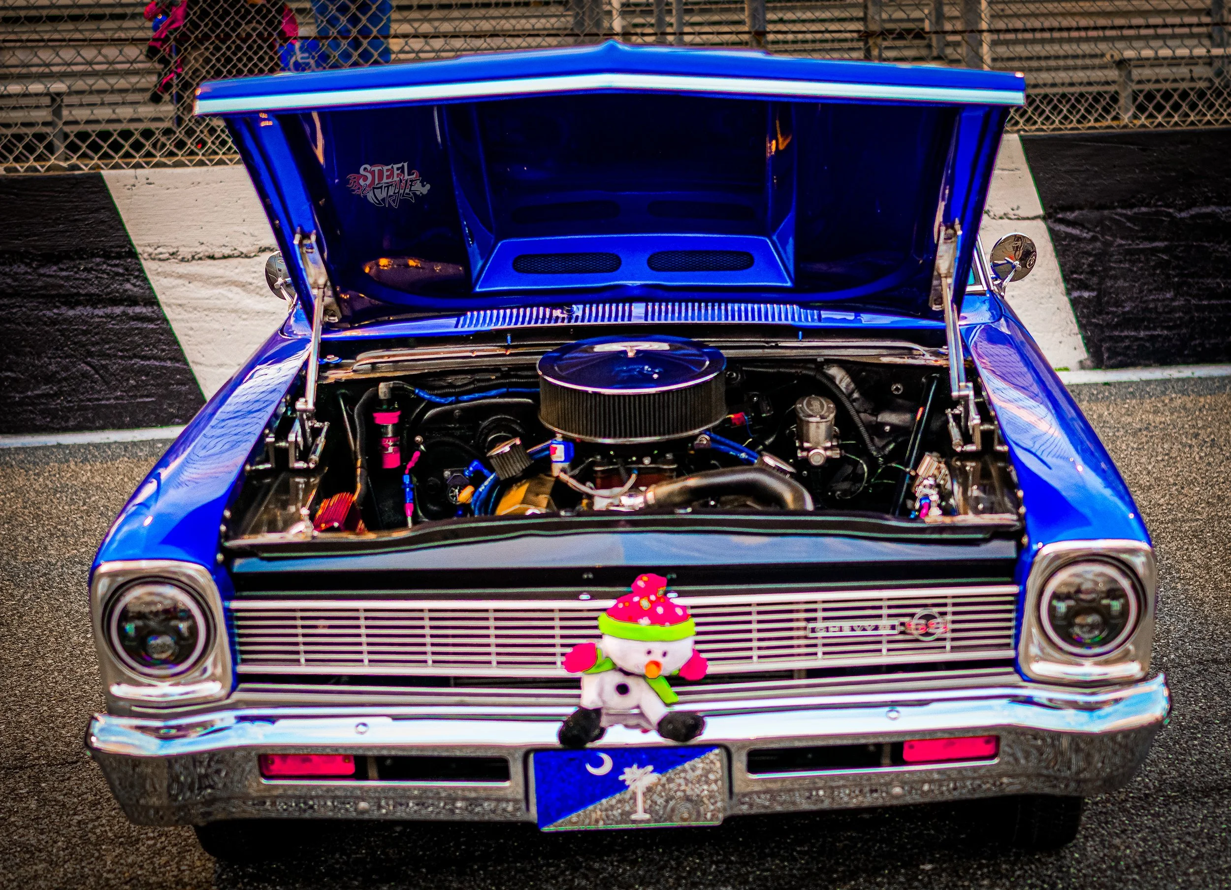 A blue classic car with its hood open on a racetrack, decorated with a plush snowman ornament on the front grille.