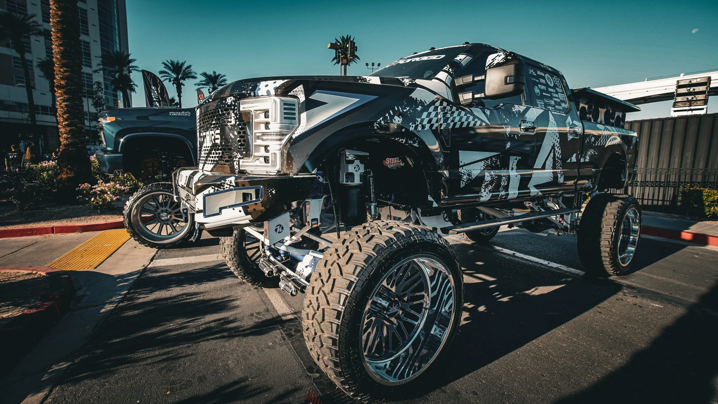 A heavily modified pickup truck with a black and white camouflage wrap, large off-road tires, and exposed suspension components parked in an outdoor lot during daytime.