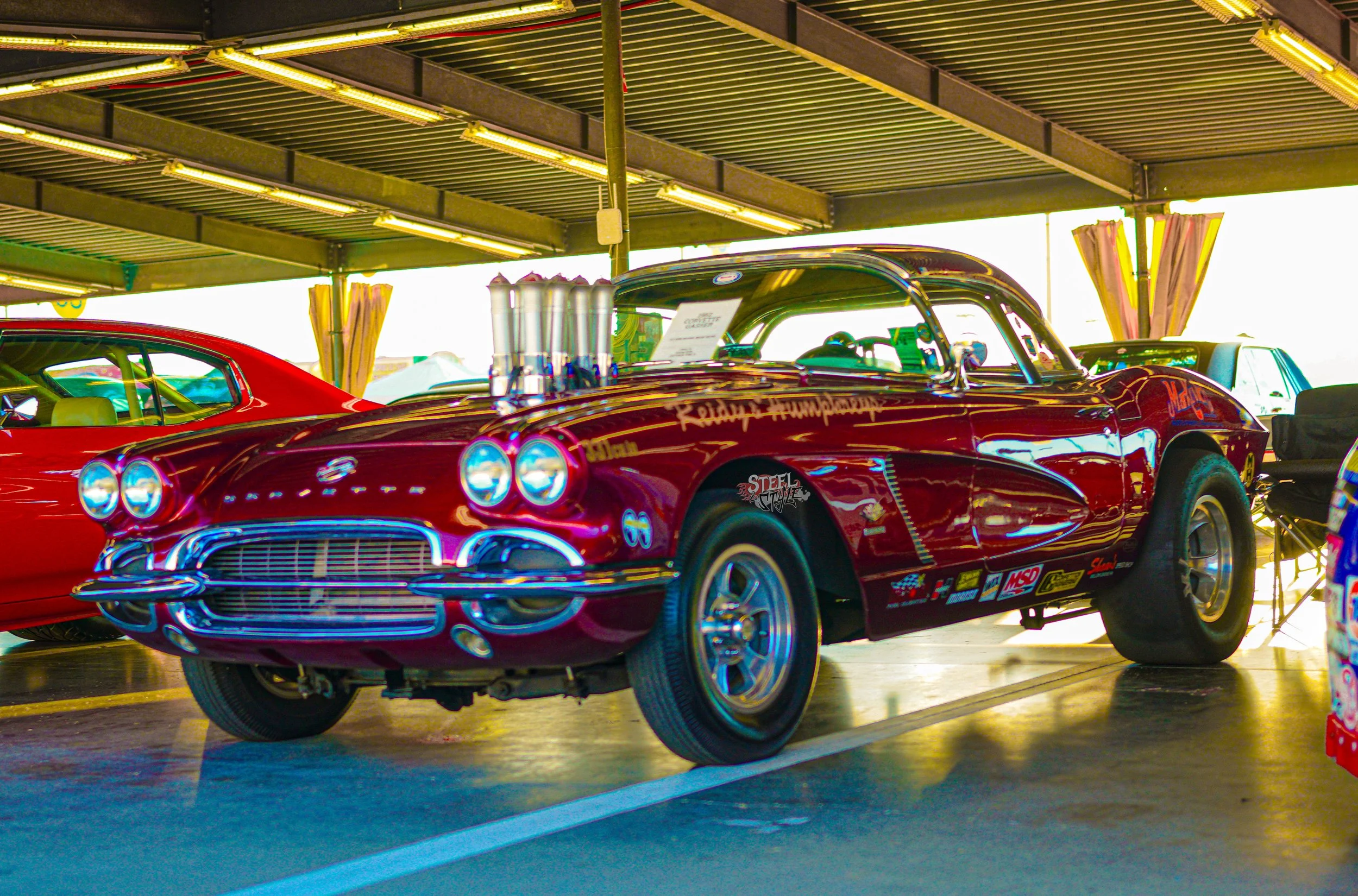 Red vintage Chevrolet Corvette race car with engine modifications, displayed in a covered parking area alongside other cars, with the front wheels turned slightly to the left.
