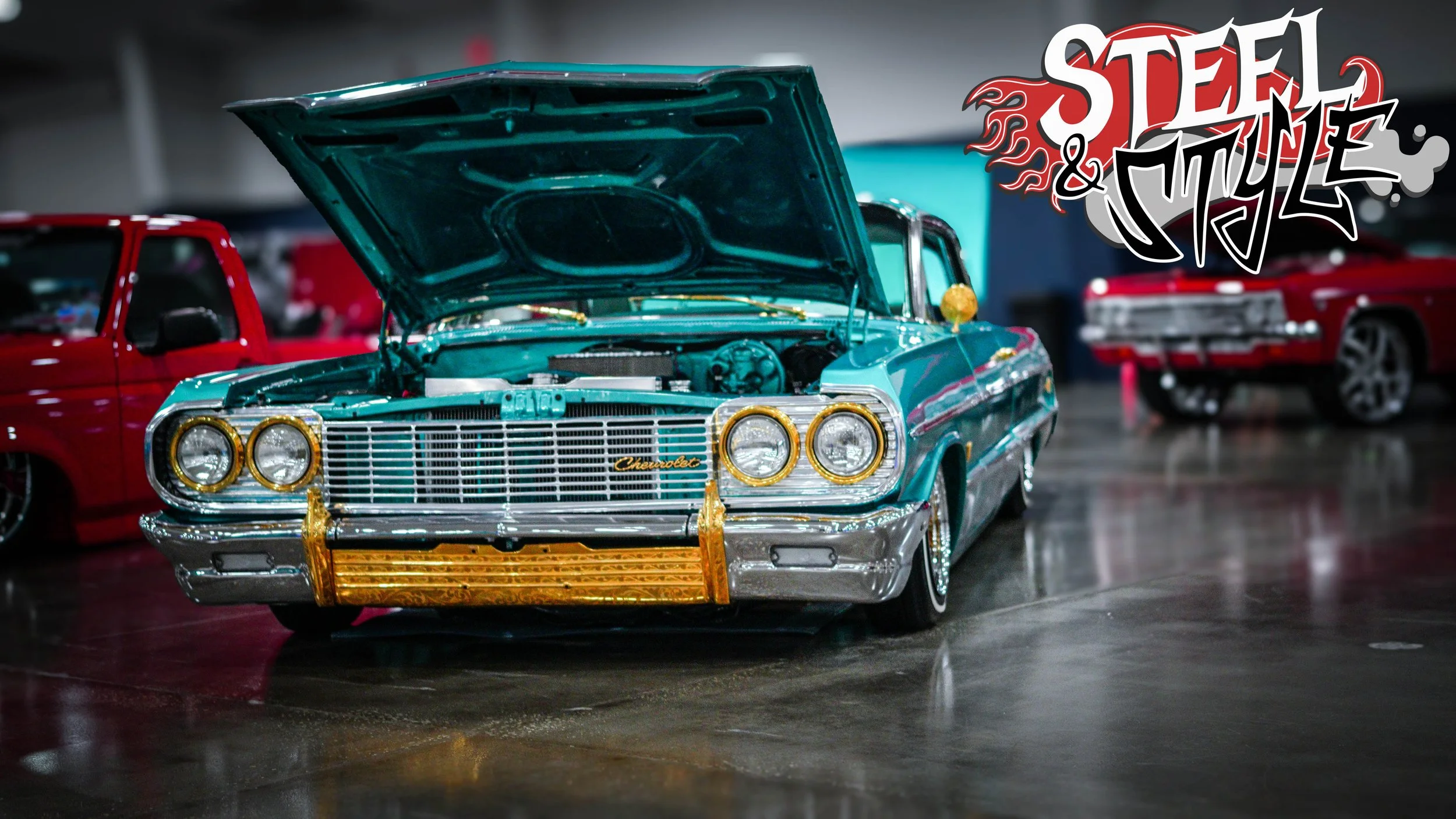 Vintage turquoise Chevrolet car with its hood up, displayed at an indoor car show, with a red car in the background and a 'Steel & Style' graphic overlay.