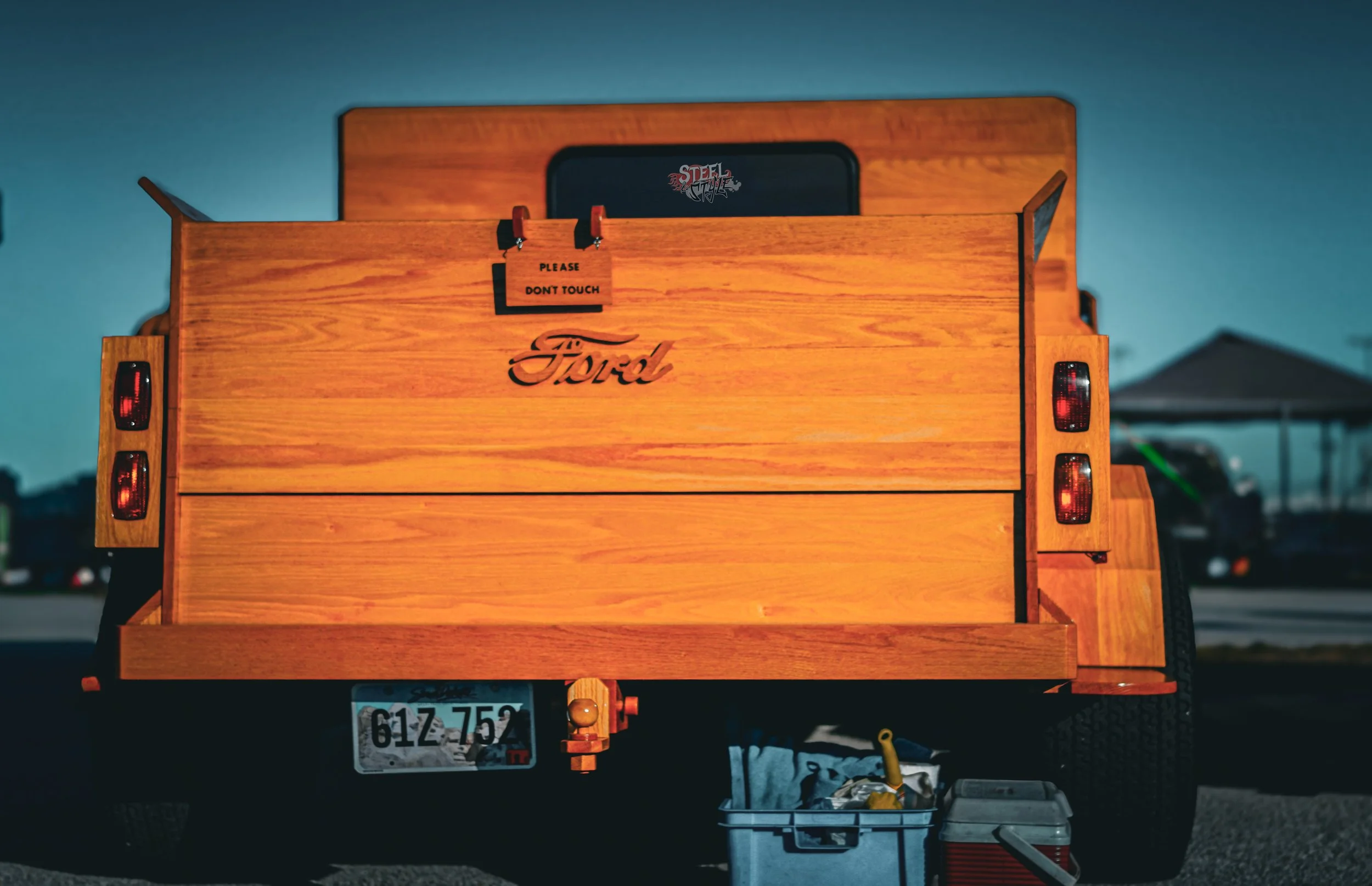 Back of a pickup truck with a wooden cargo bed, displaying a Ford emblem, taillights, and a sign that reads 'Please Don't Touch'. There are tools and containers on the ground underneath.