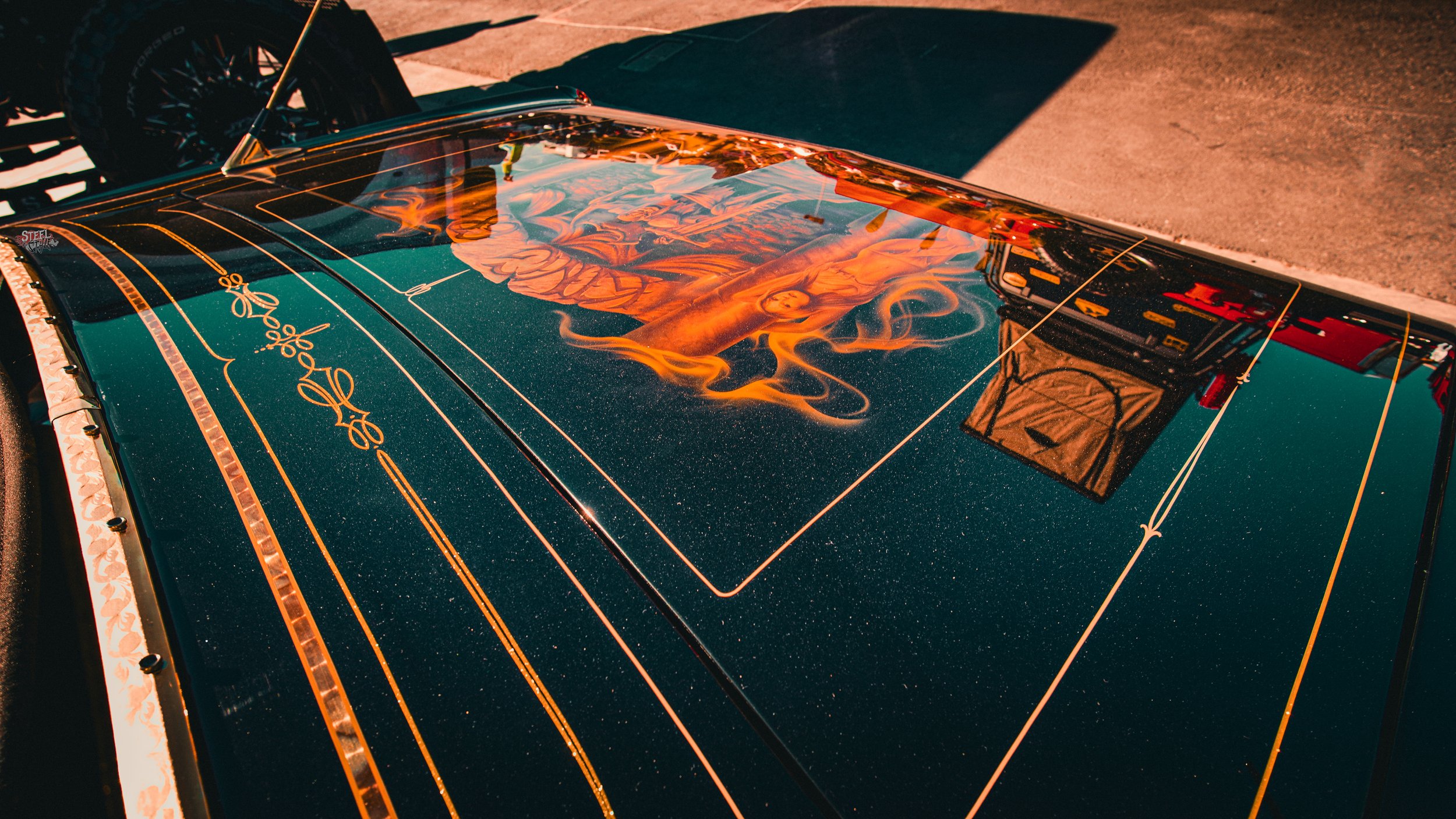 Close-up of the hood of a classic car with a detailed painting of a woman with long hair and flames, featuring gold and orange accents and reflections of a nearby structure.