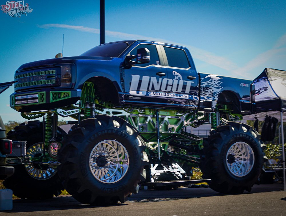 A large monster truck on display with an enclosed cab, elevated on oversized tires, green suspension components, and racing decals, parked outdoors under a blue sky.