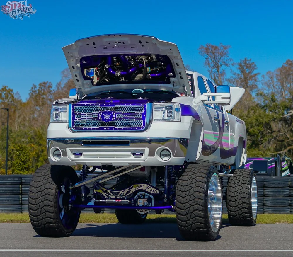 A lifted white and purple custom truck with large tires, modified front grille, and an exposed engine under the open hood, parked outdoors on a racing track.