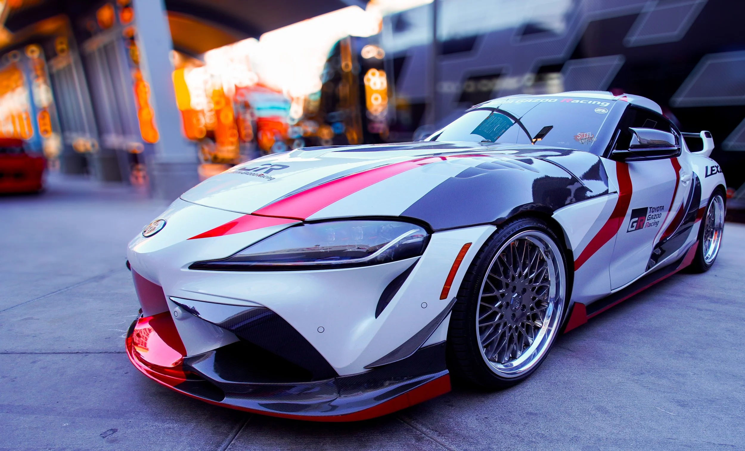 A white racing sports car with red and black accents, parked on an urban street with colorful blurred storefronts in the background.