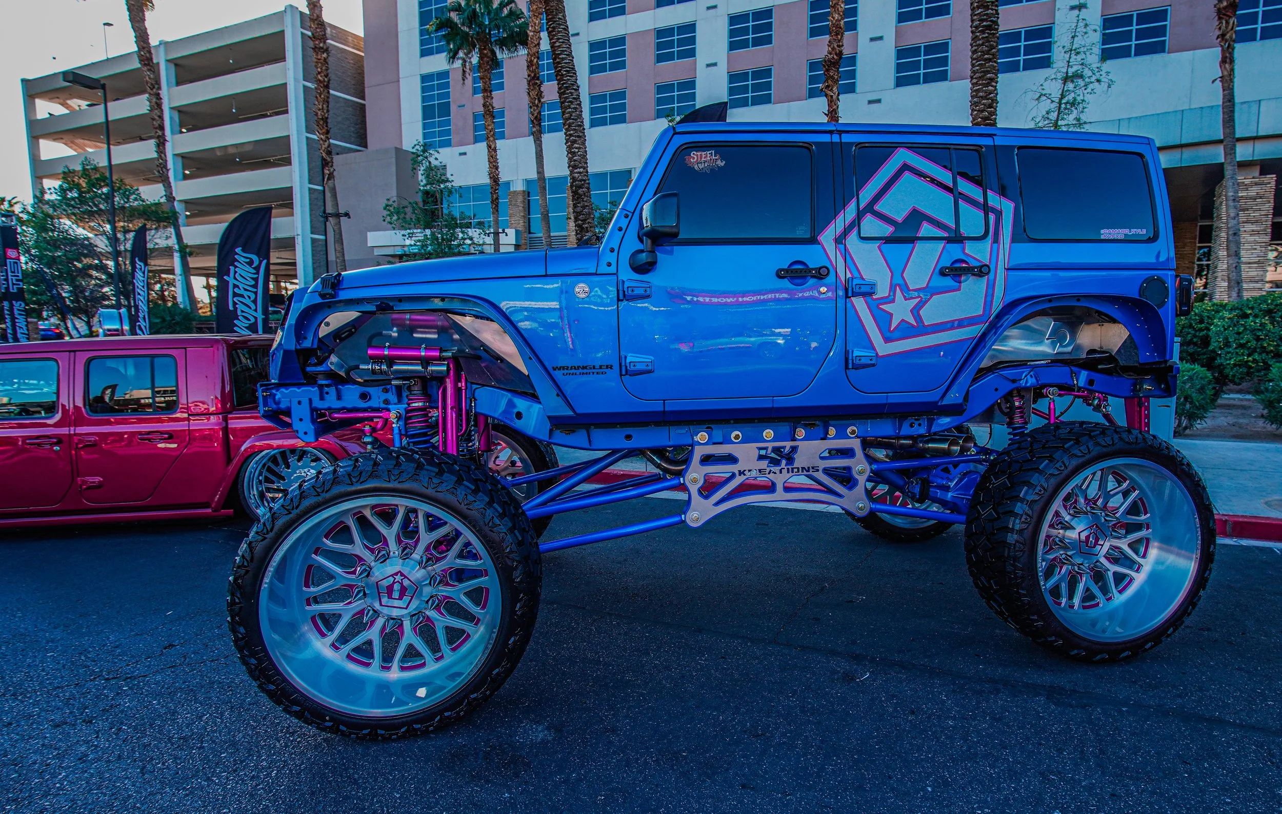 Blue customized Jeep Wrangler with large off-road tires, exposed suspension, and a distinctive star logo on the door, parked outdoors at an event with a pink vehicle and tall buildings in the background.