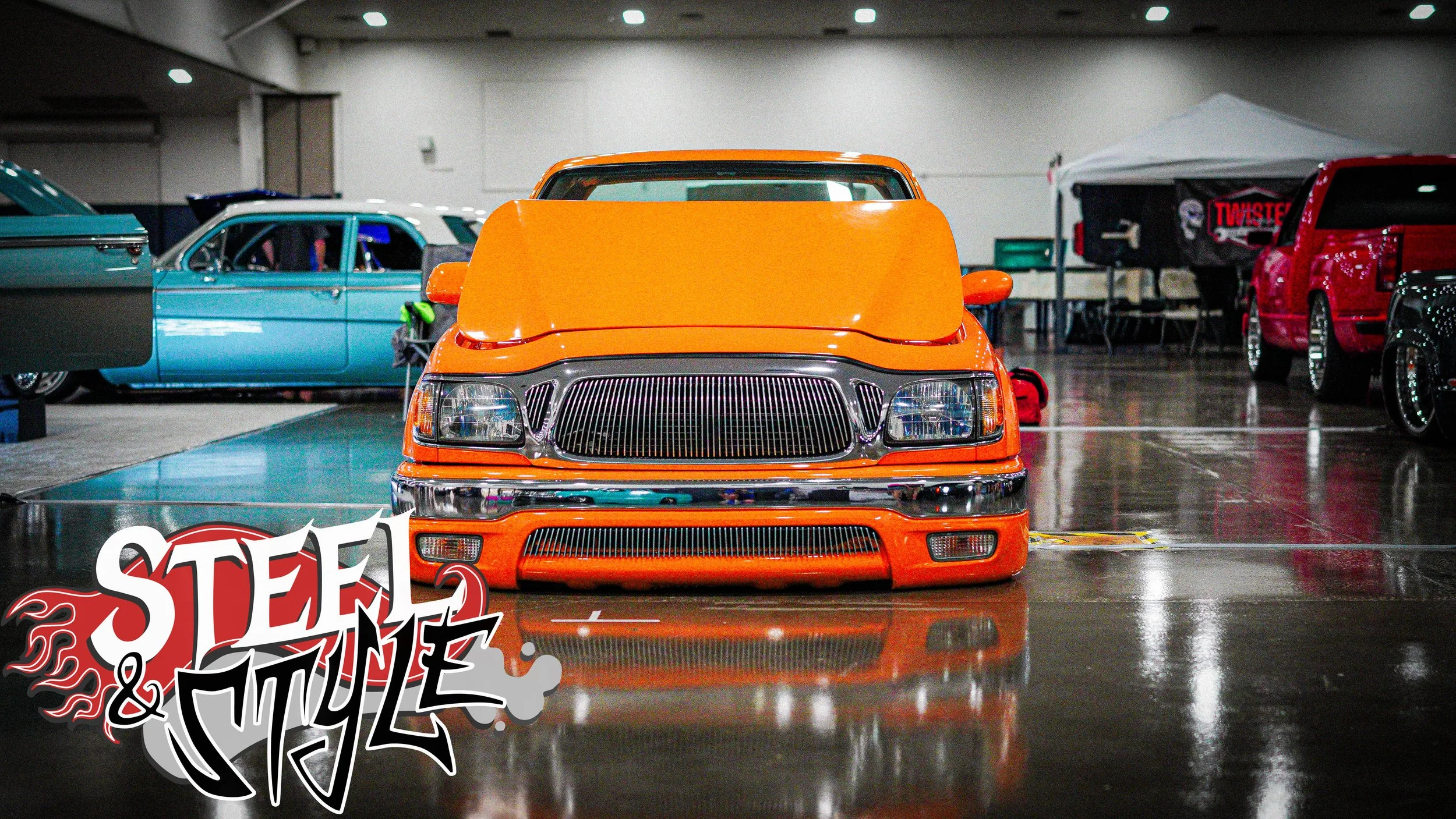 Front view of an orange lowered custom car with distinctive grill, parked indoors at a car show, with other classic cars and a tent in the background.