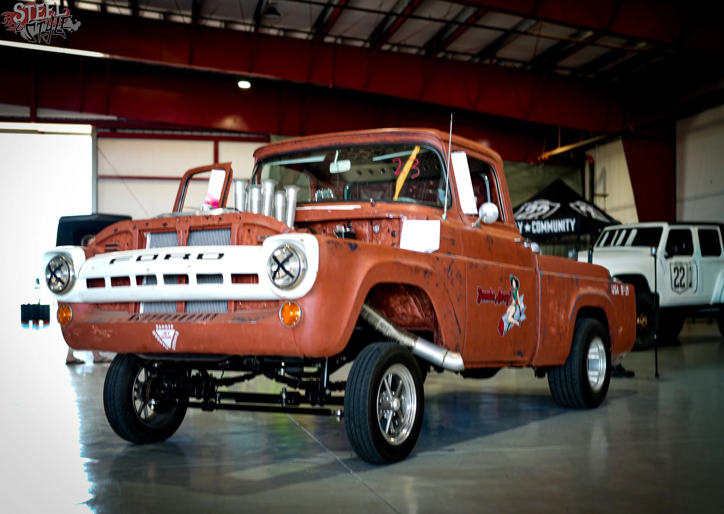 A vintage orange pickup truck with a rusty finish displayed indoors, with modifications that include raised suspension, large tires, and visible four exhaust pipes on the hood. There are some stickers on the front bumper and side of the truck. In the