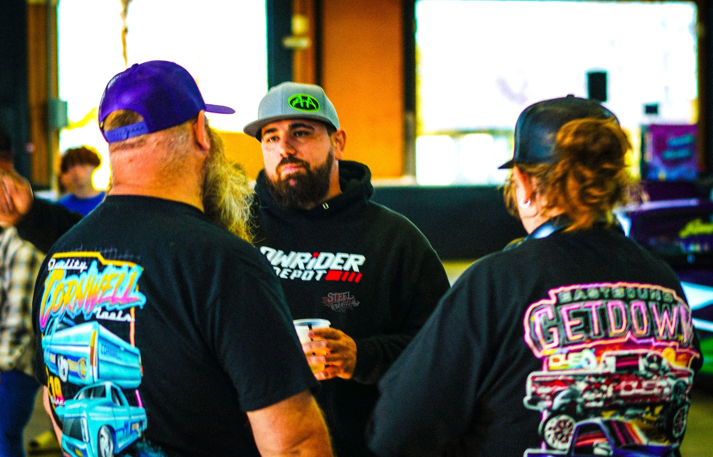Group of three people talking indoors, wearing black hoodies and caps, with colorful car-themed designs on their clothing.