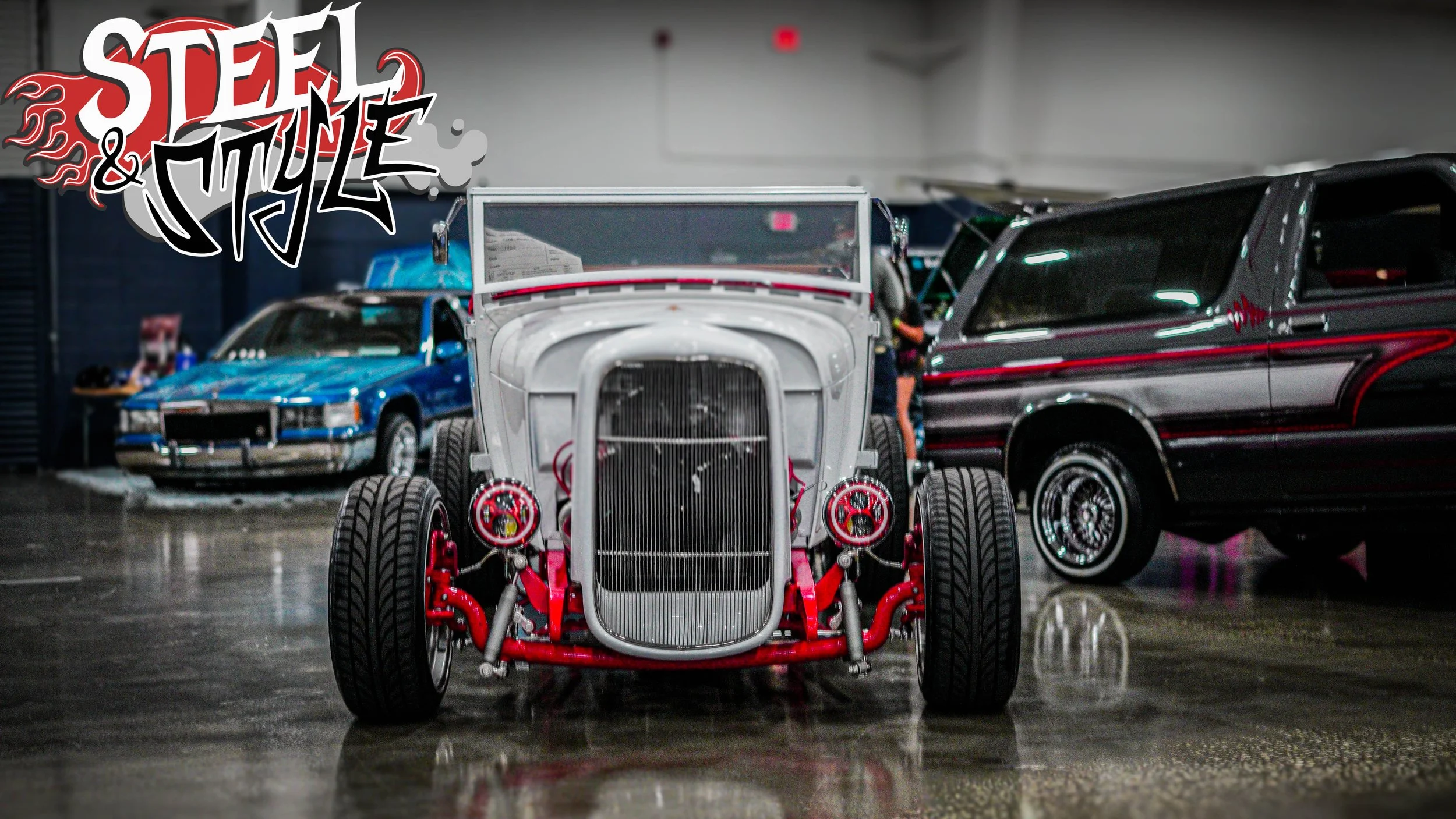 Vintage race car with exposed red chassis and large grille, displayed indoors with classic cars in background, at an event called 'Steel & Style'.